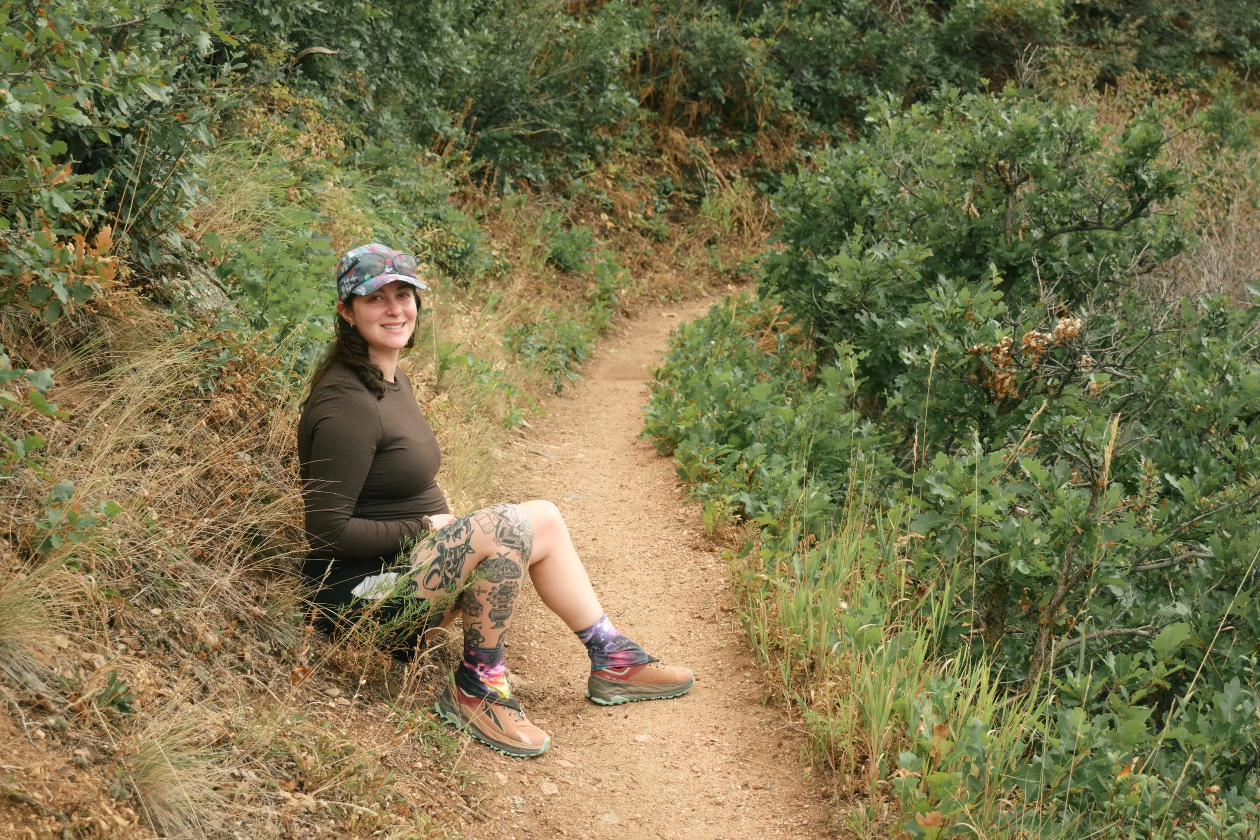 A woman with colorful tattoos on her legs, wearing a black long-sleeve shirt, a camouflage cap, colorful socks, and hiking shoes, sitting on a dirt trail surrounded by green bushes and trees.