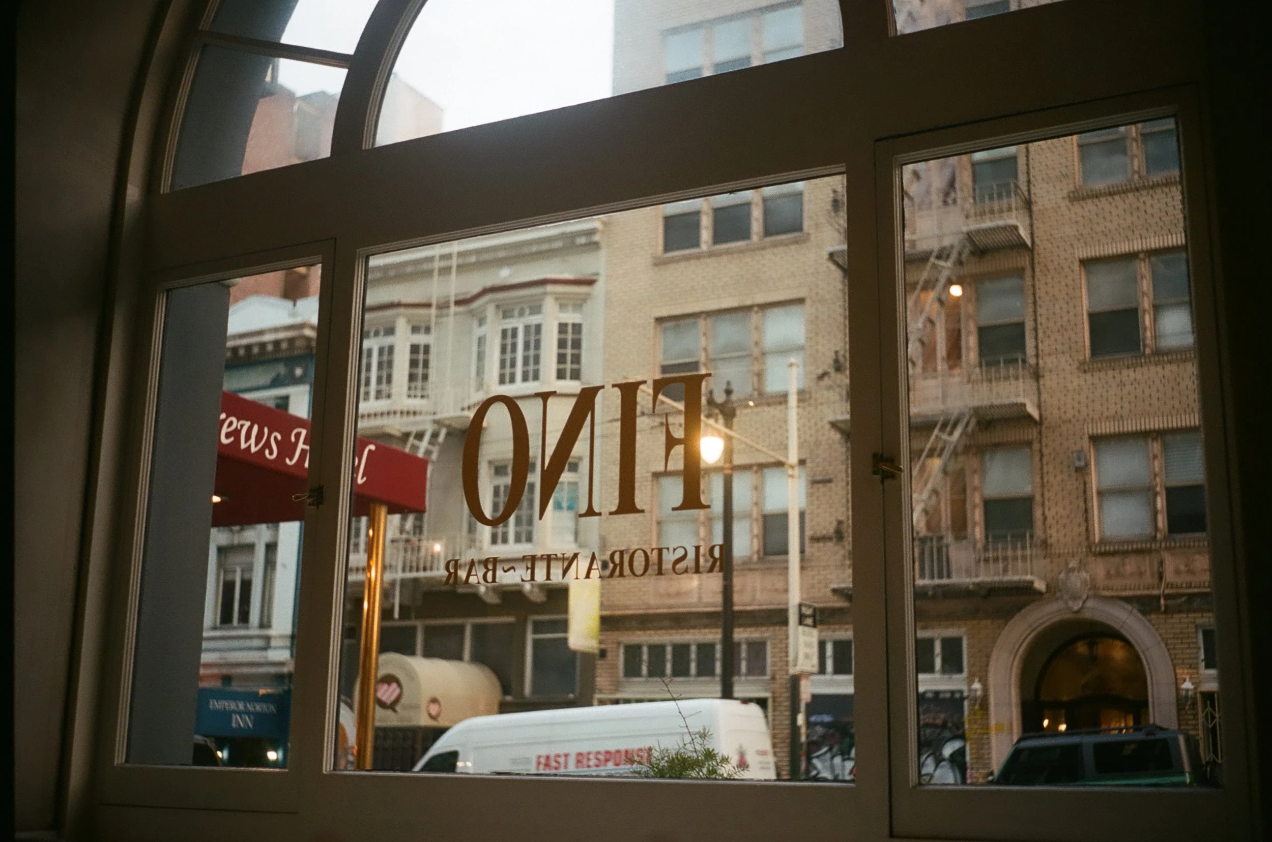 View through a window of a city street with buildings, a curbside, and a white van; the window has the words 'FINO' and 'RISTORANTE - BAR' written on it.