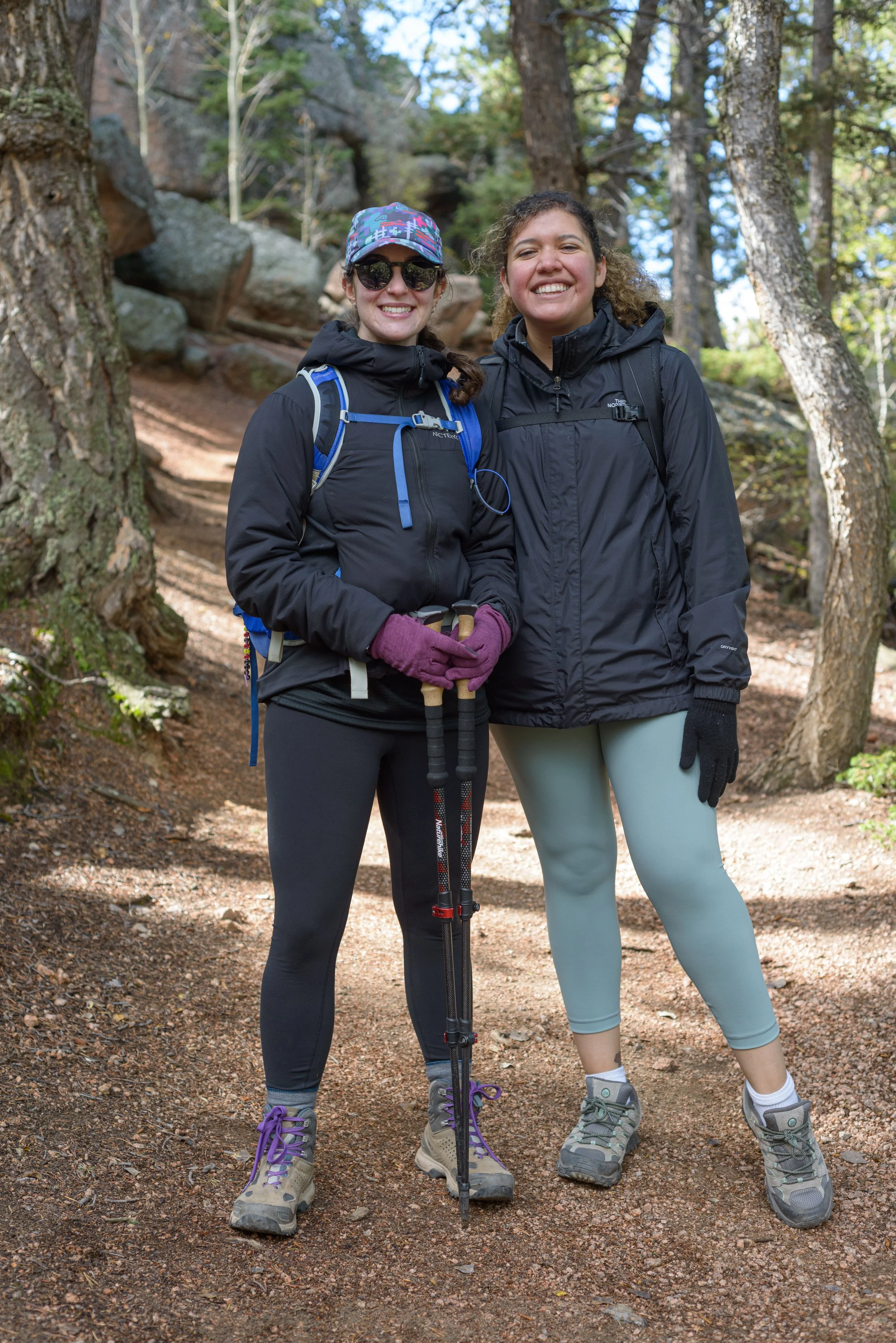 Two women in outdoor hiking gear smiling on a forest trail.
