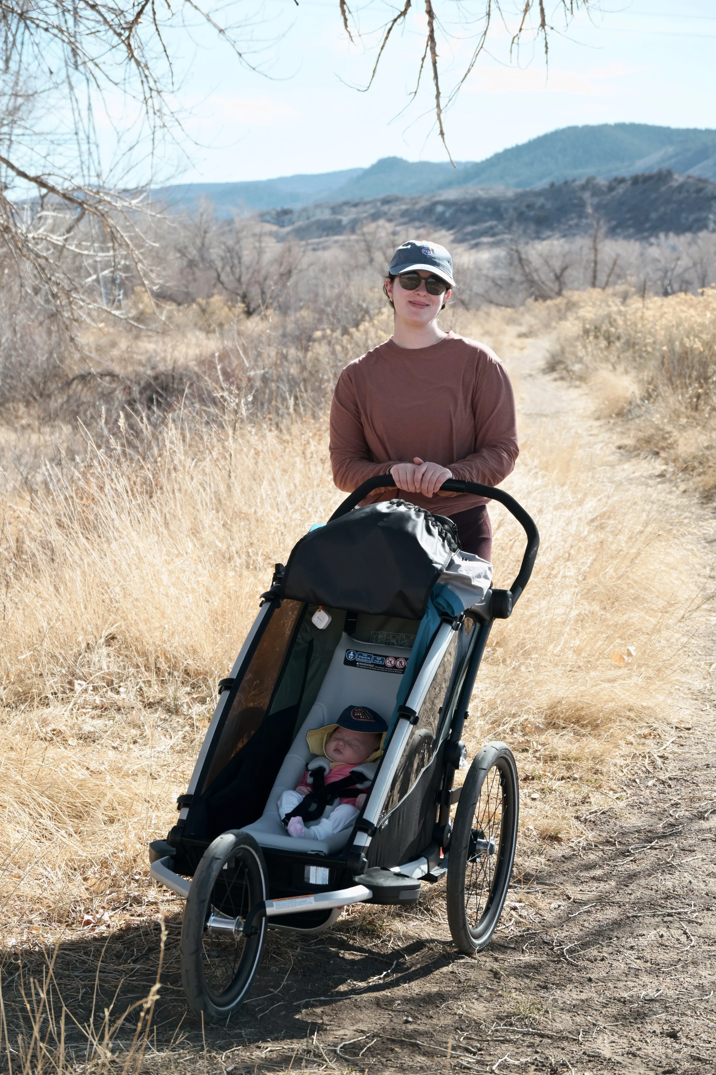 A woman with sunglasses and a baseball cap pushes a stroller with a sleeping baby along a dirt trail in a dry, grassy landscape with leafless trees and mountains in the background on a sunny day.