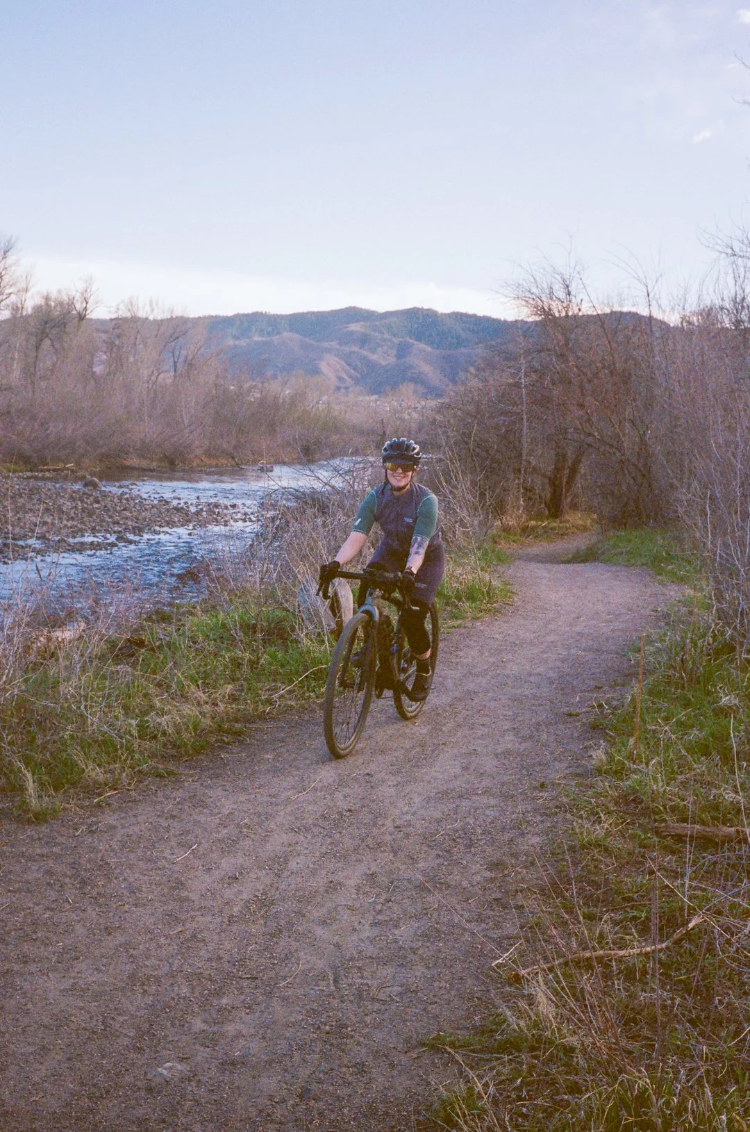 A person biking on a dirt trail near a river with mountains in the background.