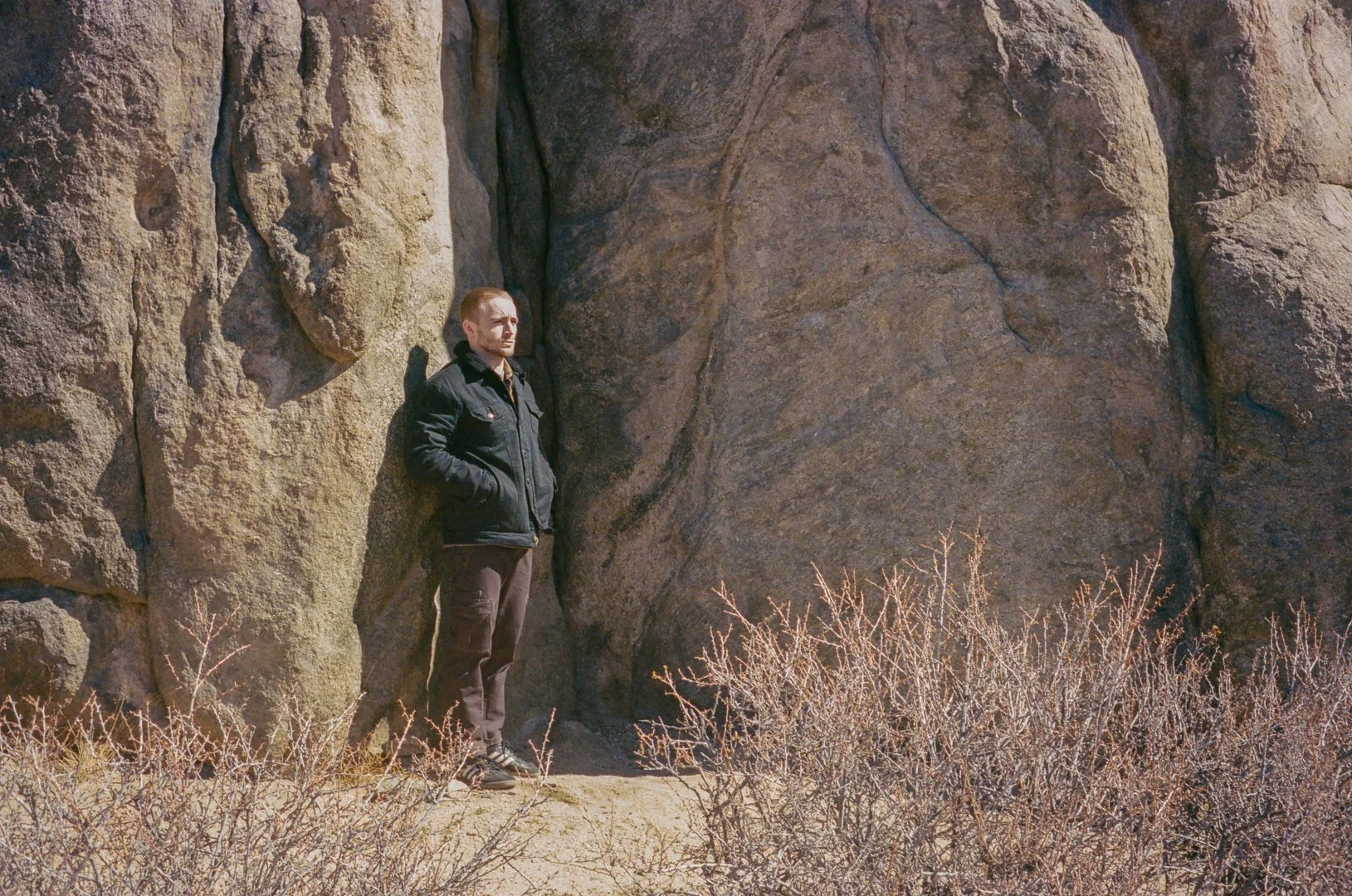 A man wearing a black jacket and brown pants standing next to a large rock formation with dry bushes in front.