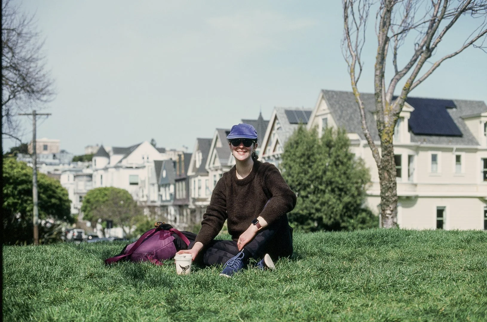 A woman sitting on green grass in an urban park, smiling, wearing sunglasses, a blue hat, dark sweater, and sneakers, with a backpack and a cup nearby, residential houses in the background, and a leafless tree to her right.