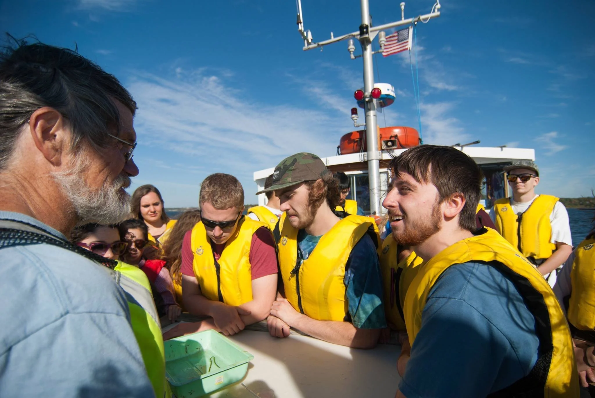 A group of people on a boat wearing yellow life jackets, listening attentively to an older man with a beard and glasses, possibly a guide or instructor, during daytime with blue sky and water in the background.