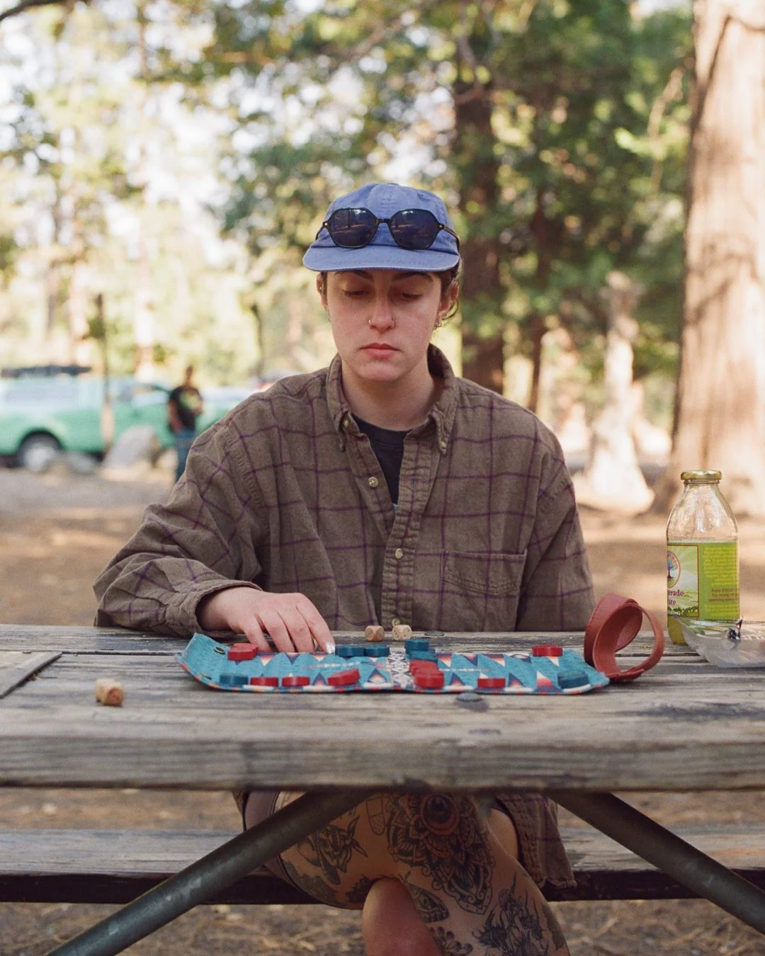A young woman with short hair, wearing a blue cap and sunglasses on top, playing a board game at a wooden picnic table outdoors in a wooded park.