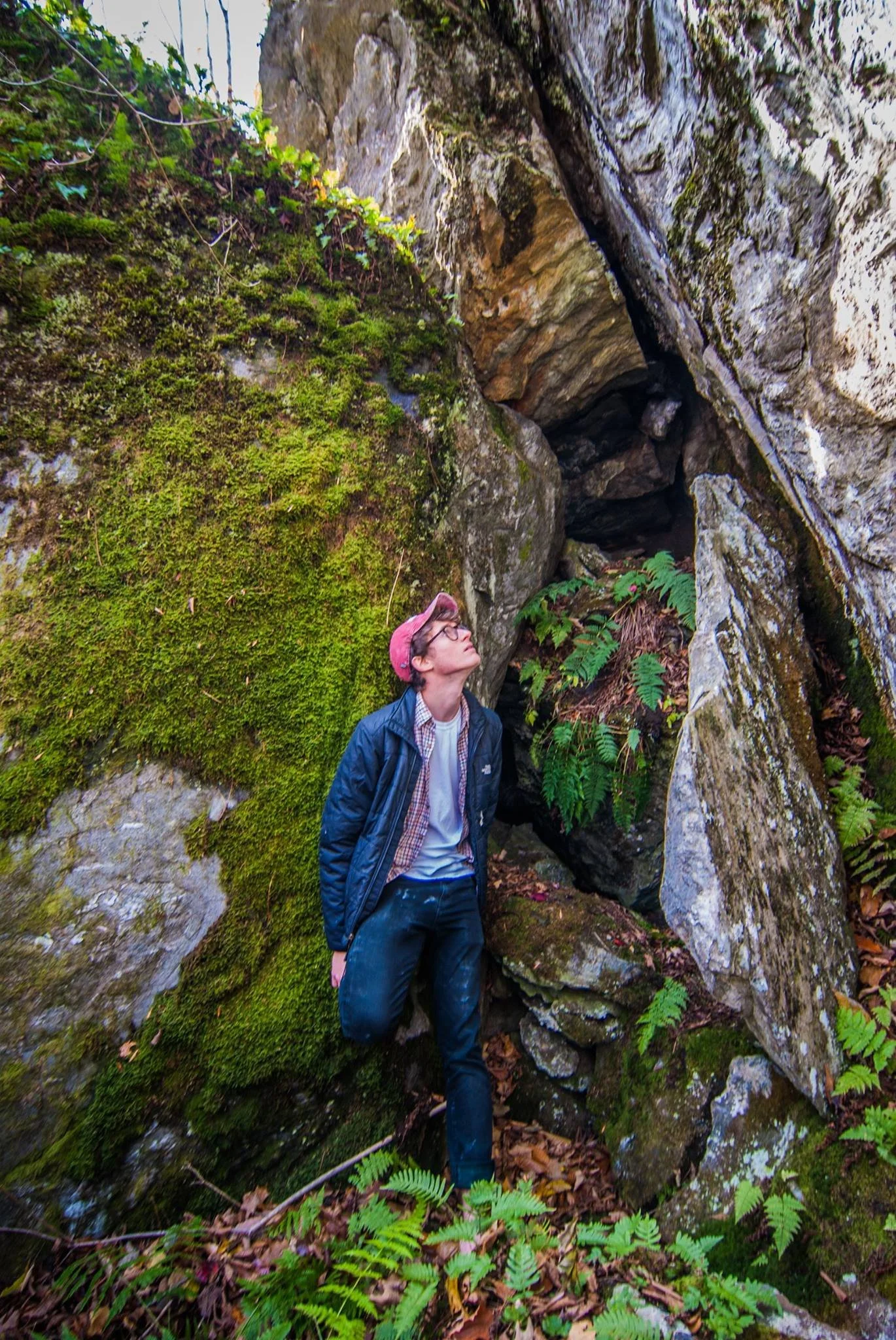 A person in a blue jacket, glasses, and a pink cap standing on a mossy rock in a forest, looking up at large rocks and ferns.
