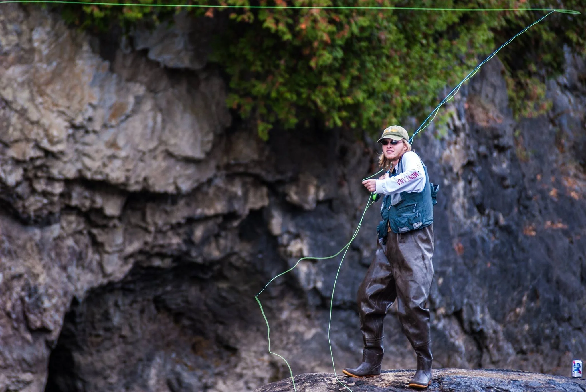A woman standing on a rock near a rocky shoreline, holding a green fishing line with her right hand. She is wearing a white long-sleeve shirt, a black life vest, brown waterproof pants, and rubber boots. She has sunglasses, a cap, and a backpack.