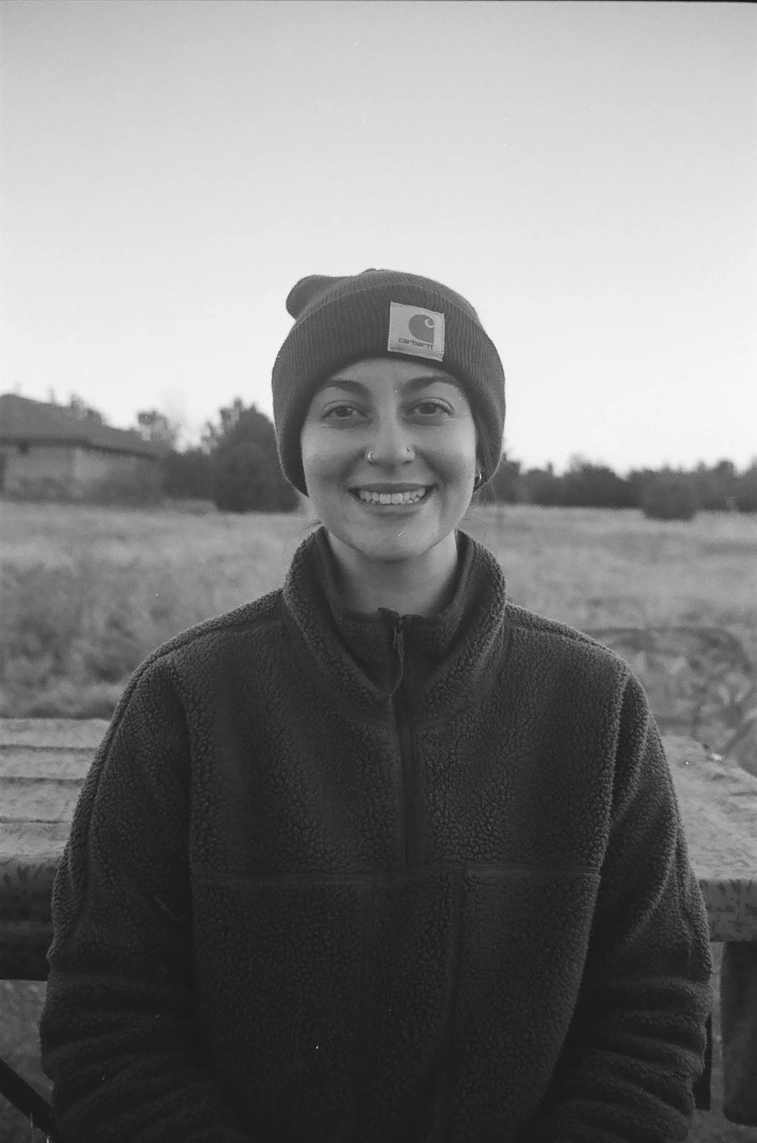 A smiling woman wearing a Carhartt beanie and a fleece jacket, sitting outdoors with an open field and houses in the background.