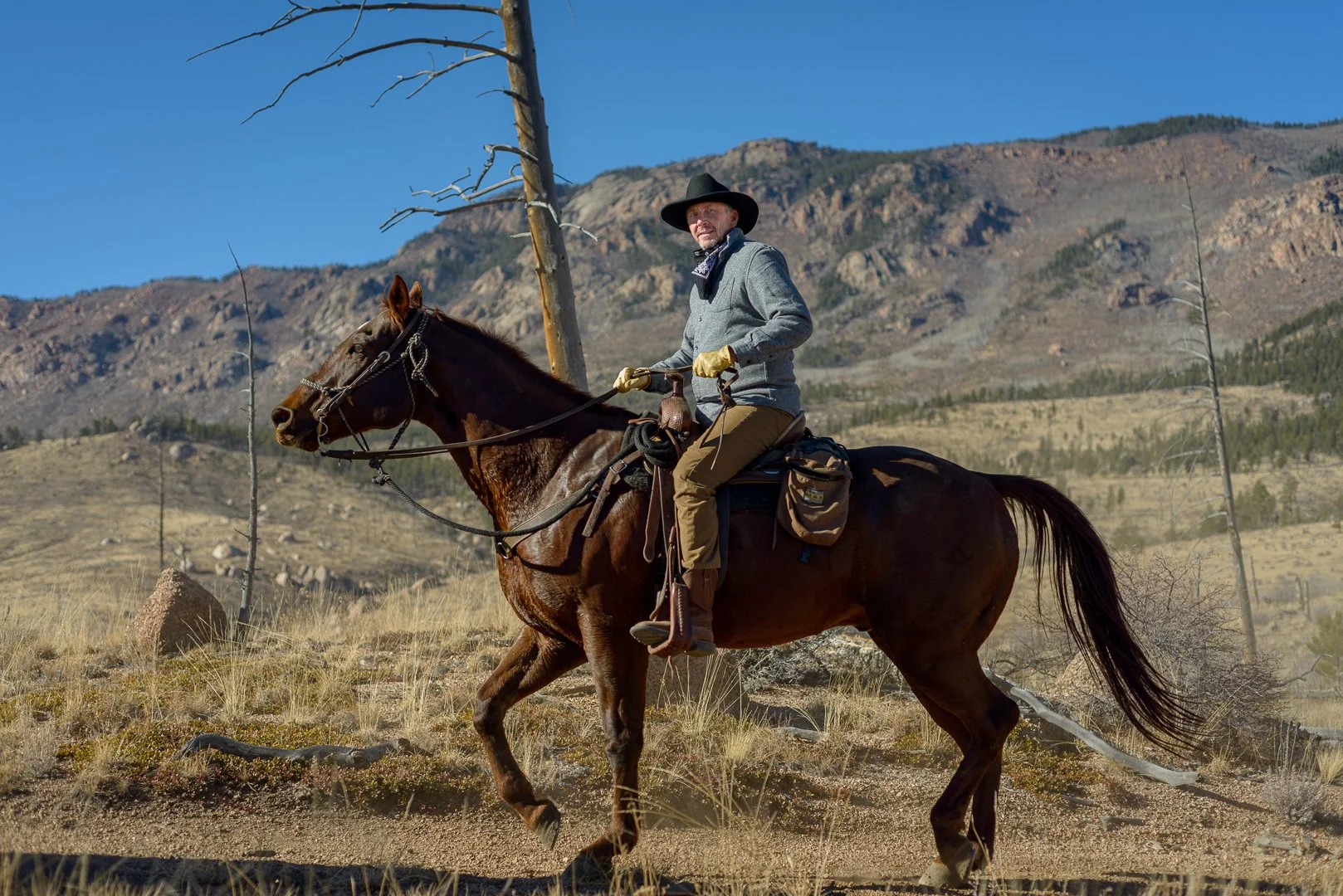 A man riding a brown horse in a dry, mountainous landscape with sparse trees and grass, under a clear blue sky.