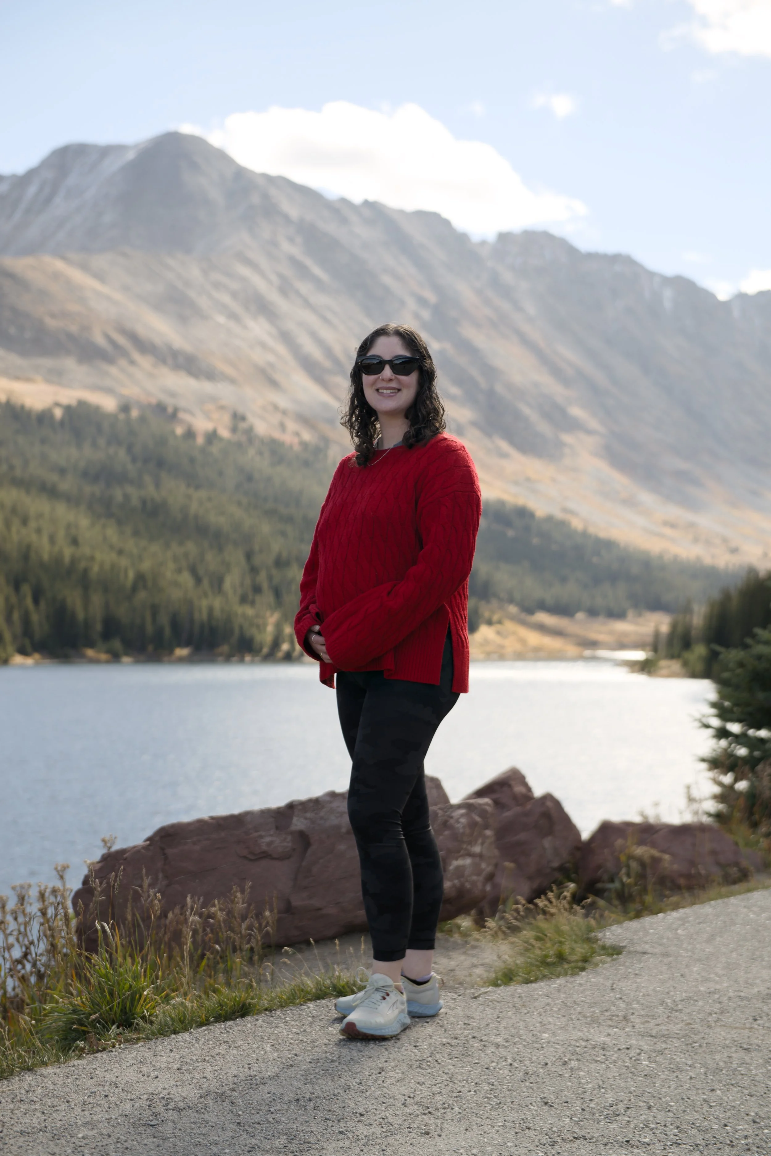 A woman wearing sunglasses and a red sweater standing on a trail beside a lake with mountains in the background.