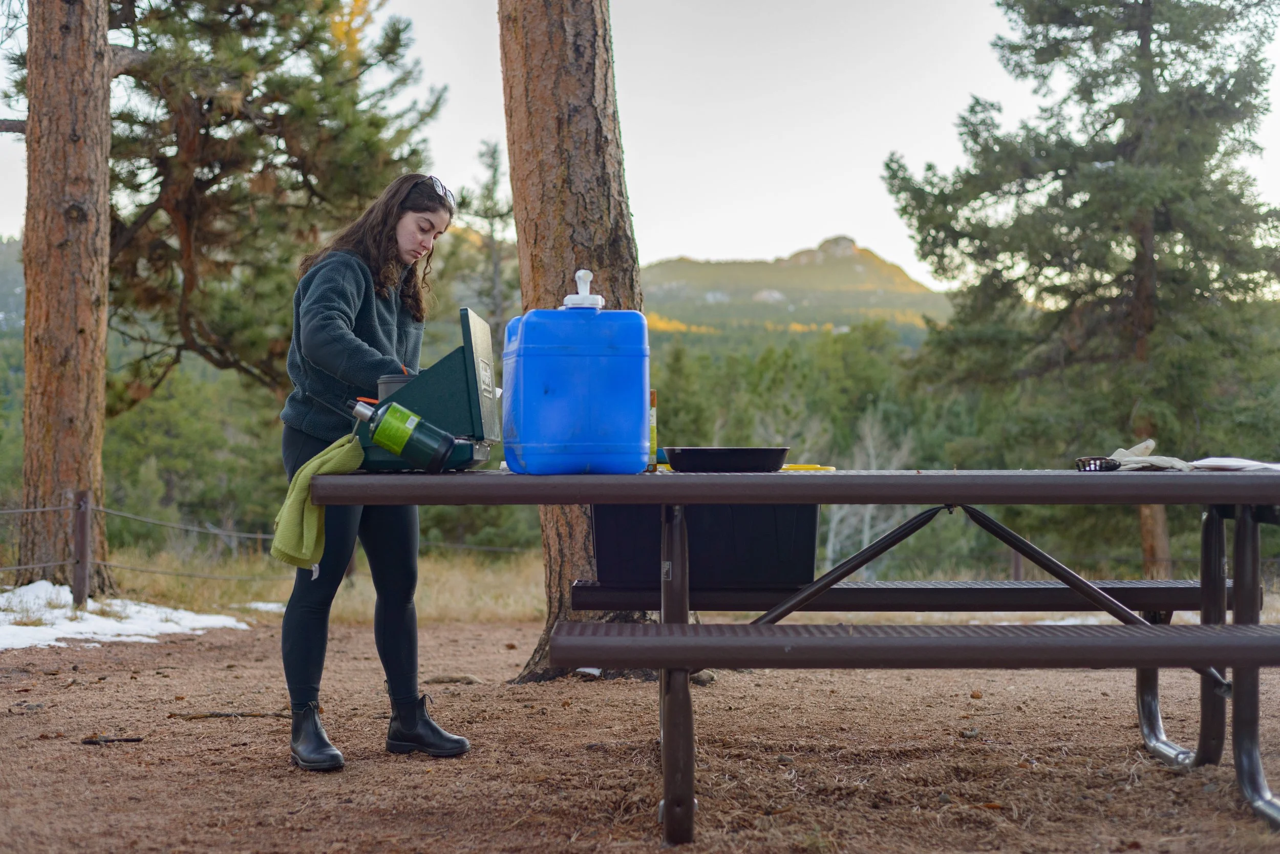 A woman in outdoor camping gear stands at a picnic table in a forested area, looking at a portable stove or small cooking device.
