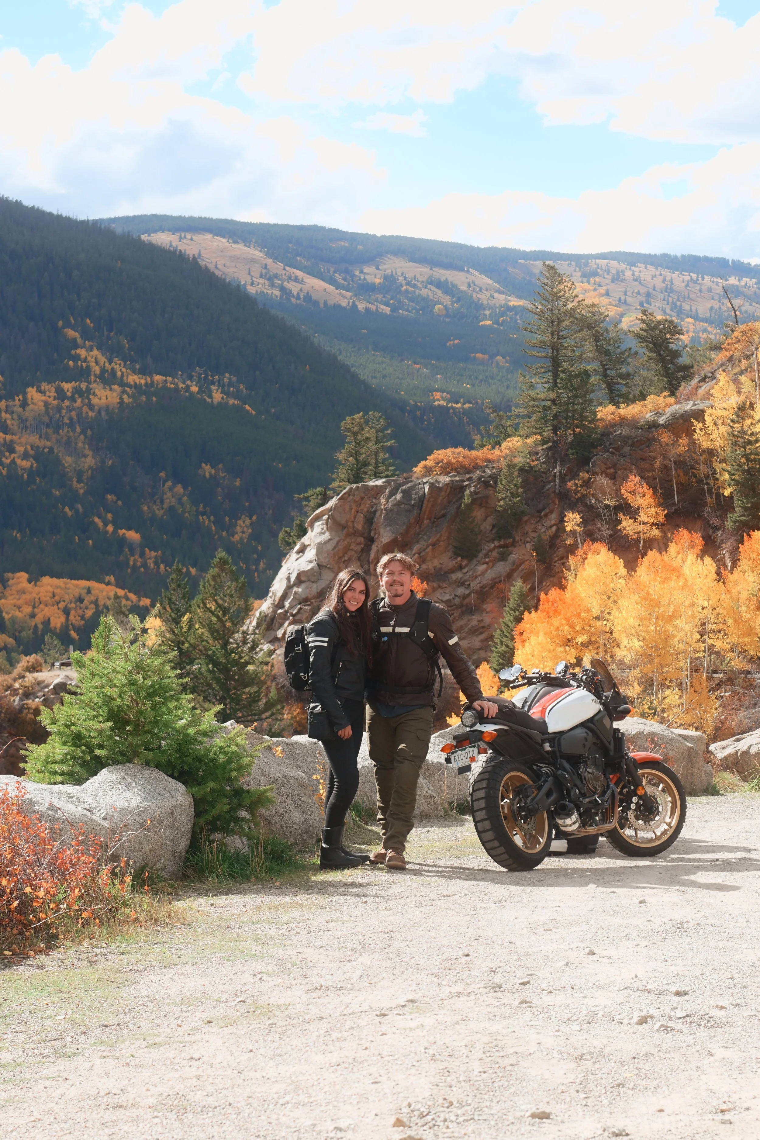 A couple stands smiling next to a motorcycle in a mountainous area with fall-colored trees.