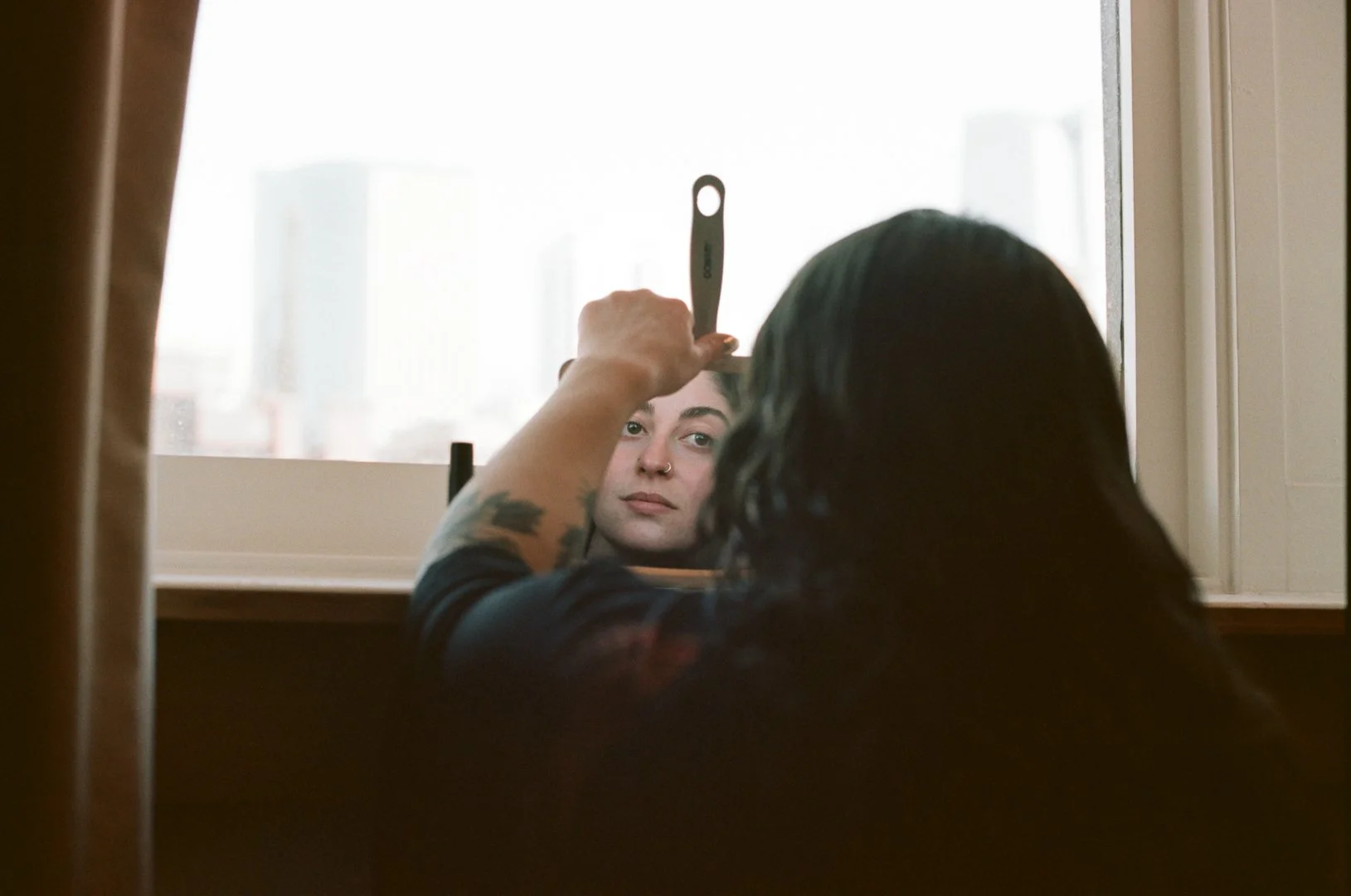A woman with dark hair looks into a mirror while a person with tattoos on their arm uses a metal tool to shape her eyebrows, with a city skyline visible through the window in the background.