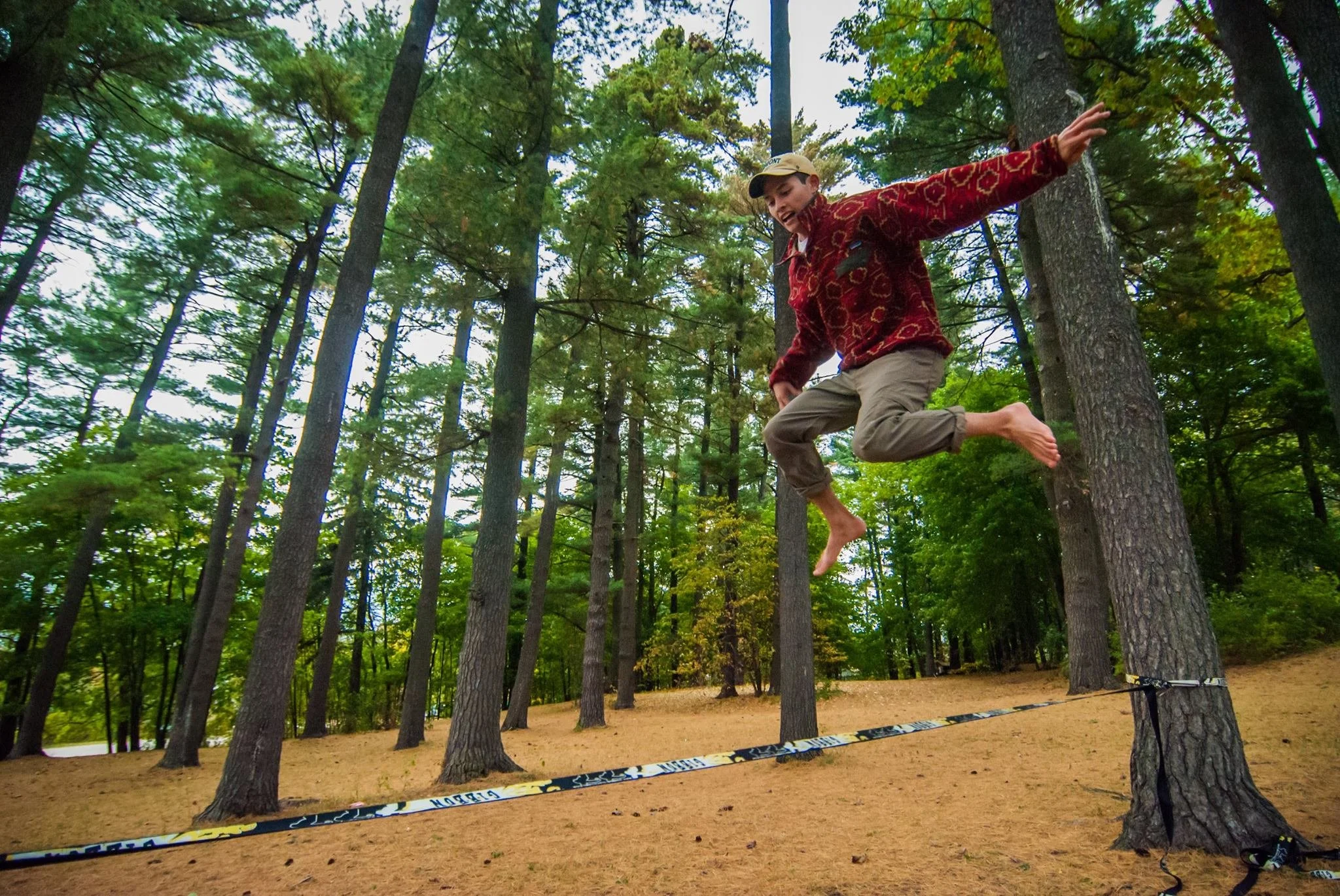 Child jumping in a forested park, balancing on a slackline tied between two trees, wearing a red patterned jacket and beige pants without shoes.