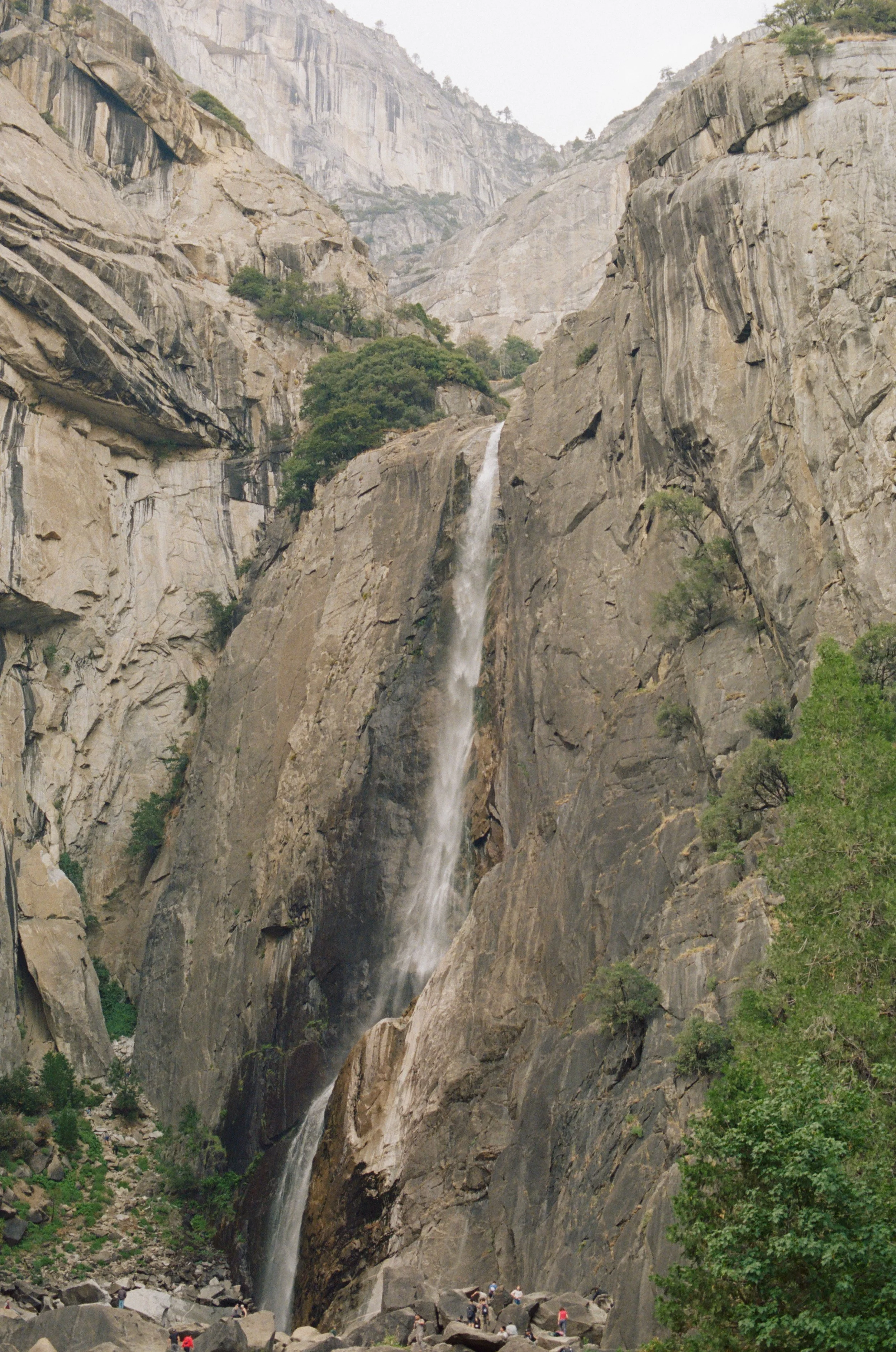 A tall, narrow waterfall flowing down a steep granite canyon with large rocks at the base, surrounded by green bushes and trees.