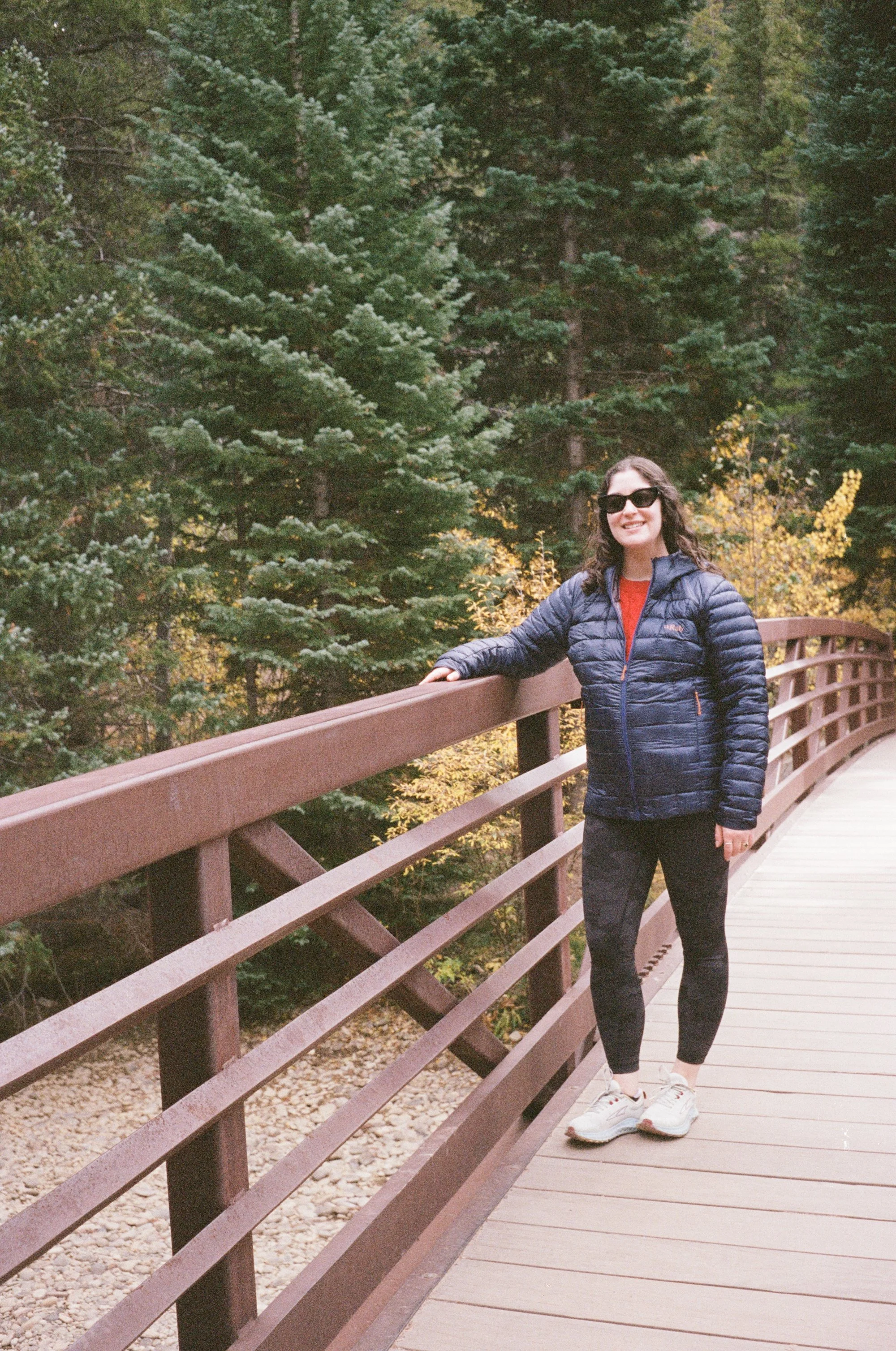 Woman standing on a wooden bridge with metal railings in a forest with evergreen trees and autumn foliage.