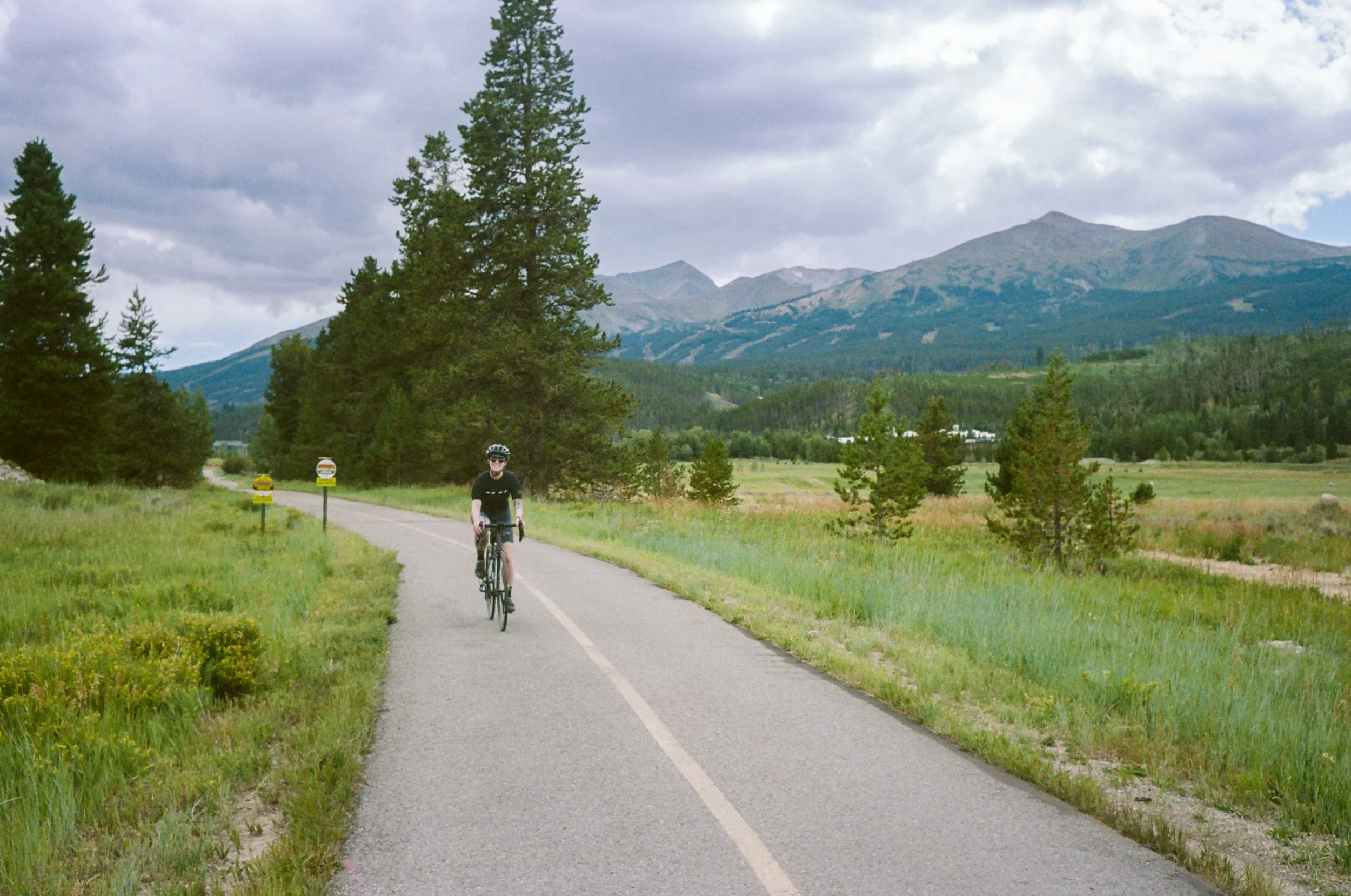 A person riding a bicycle on a paved trail through a scenic landscape with mountains, trees, and a cloudy sky.
