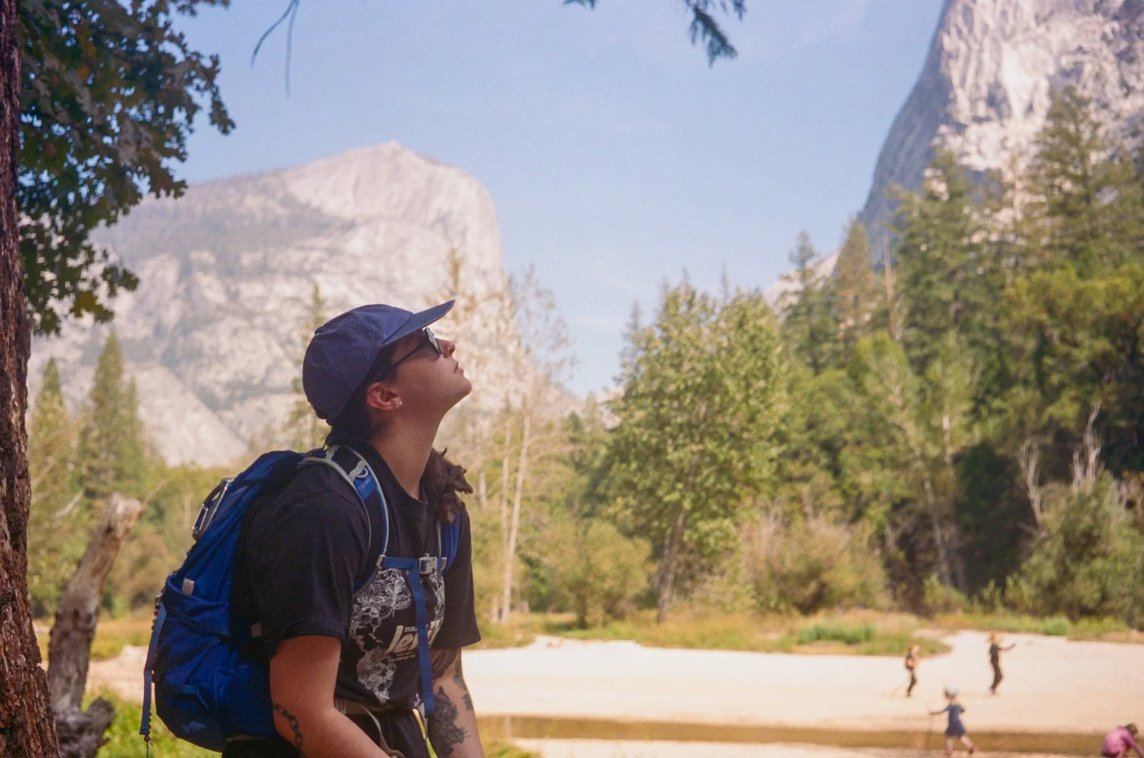 A woman hiking in a forested area with mountains in the background, wearing a hat, sunglasses, a dark shirt, and a blue backpack, looking up at the sky.