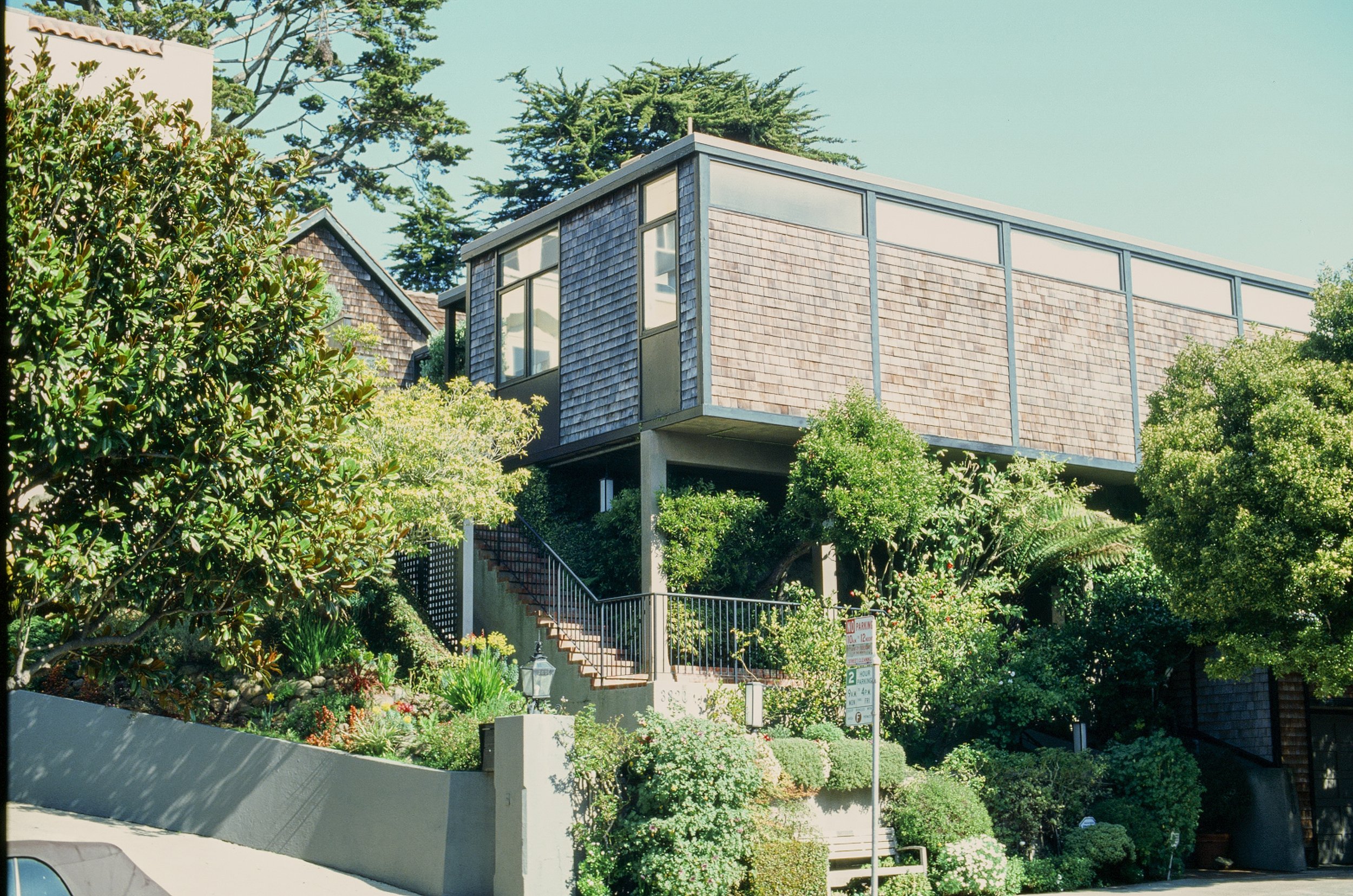 Modern house built on stilts with large glass windows, surrounded by lush greenery and trees, with a staircase leading up to the entrance.
