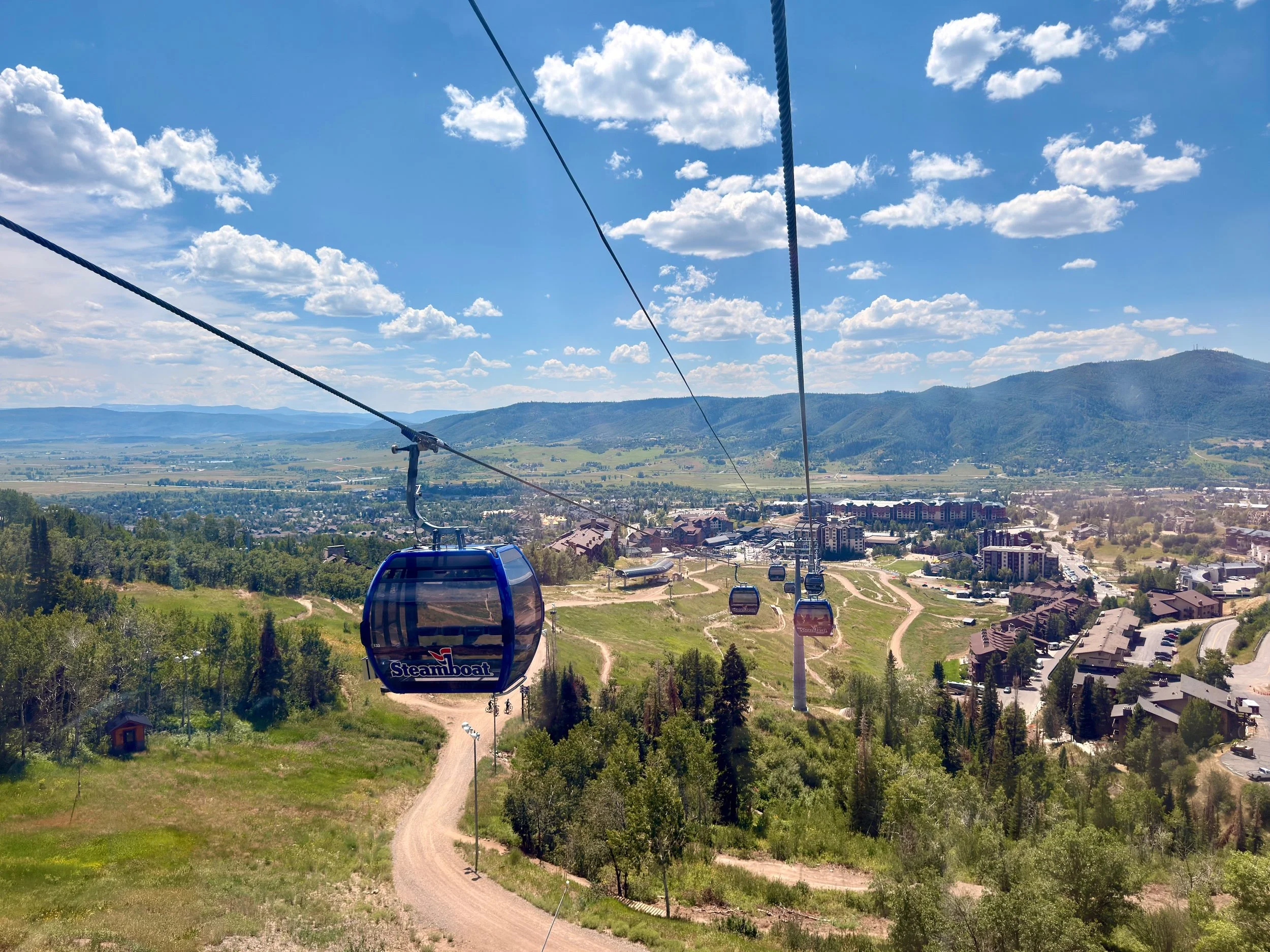 View of a sky gondola lift named Steamboat traveling over green hills and trees towards a ski resort town with mountains in the background on a sunny day with scattered clouds.