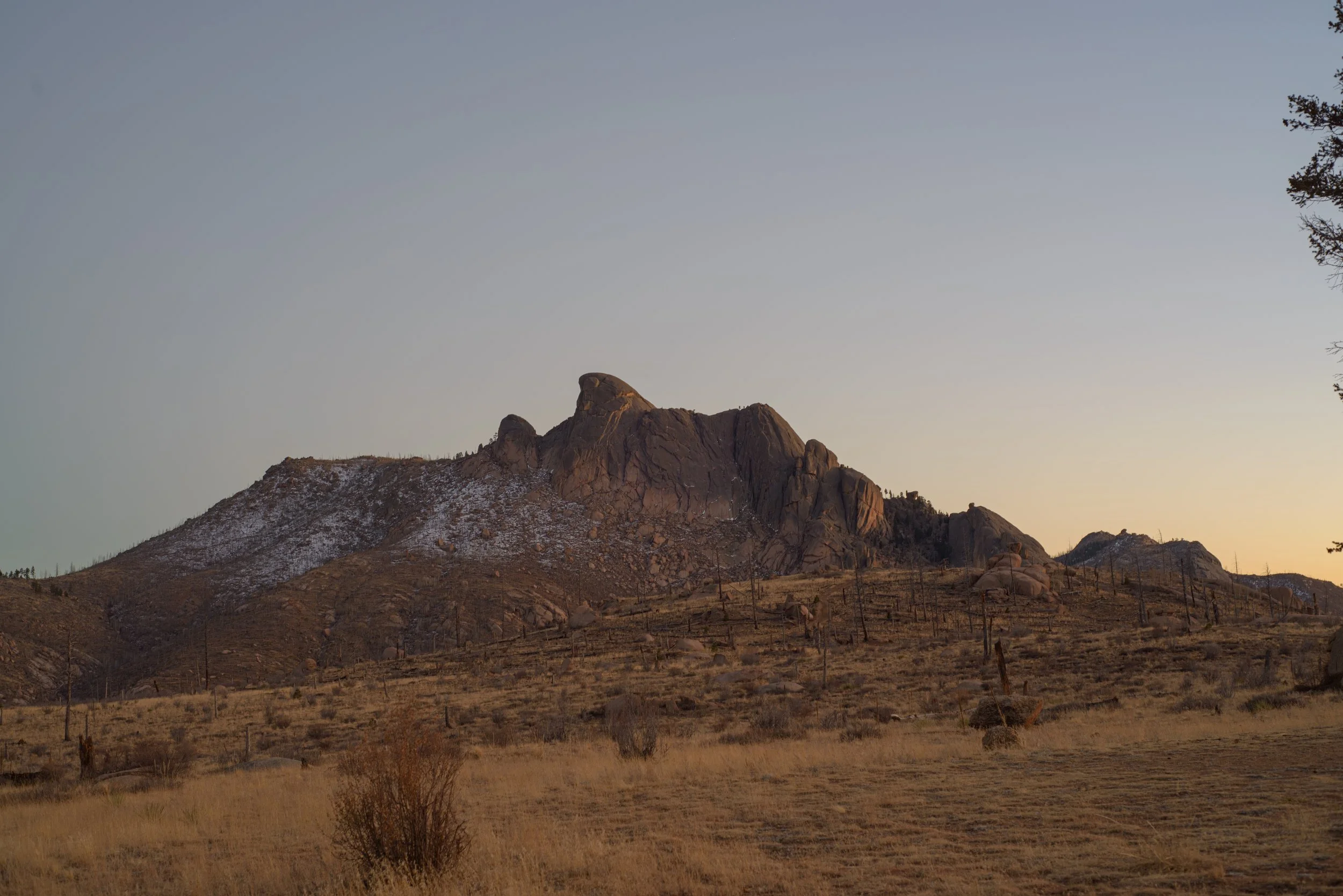 Mountain landscape with rocky peaks, some snow patches, dry grass, and sparse trees at sunset or sunrise.