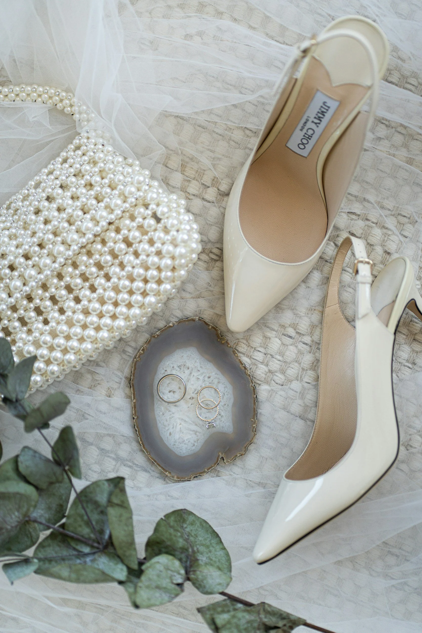 A pair of ivory high-heeled shoes, a pearl clutch, and jewelry inside a decorative tray on a textured cloth surface, with some eucalyptus leaves in the foreground.