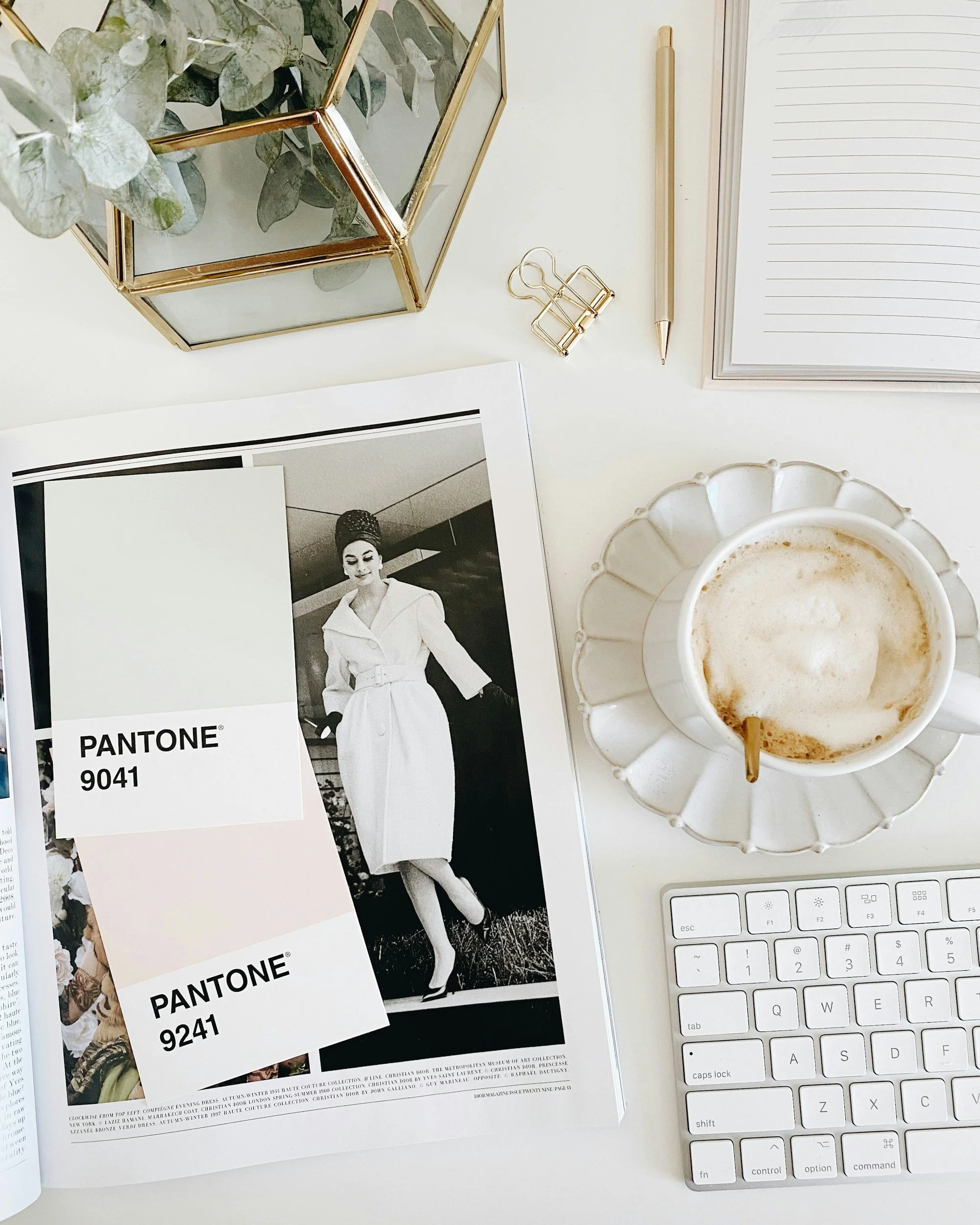 A white desk with a cup of coffee, a computer keyboard, a gold paperclip, a gold binder clip, a gold pen, and a magazine with black and white fashion photography and Pantone color swatches.