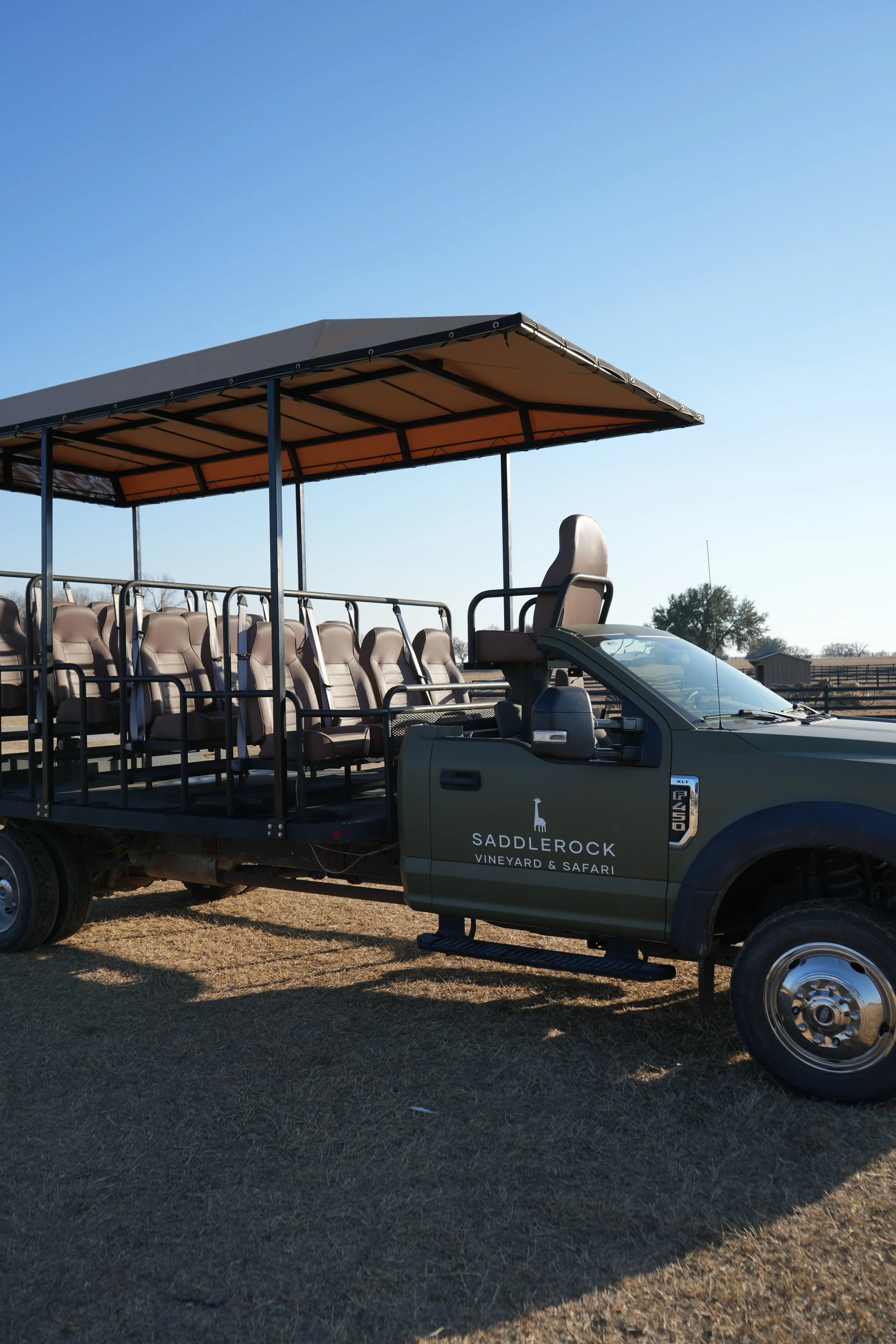 A safari vehicle with a covered seating area and a row of beige seats, parked on grass under a clear blue sky, with trees and fencing in the background.