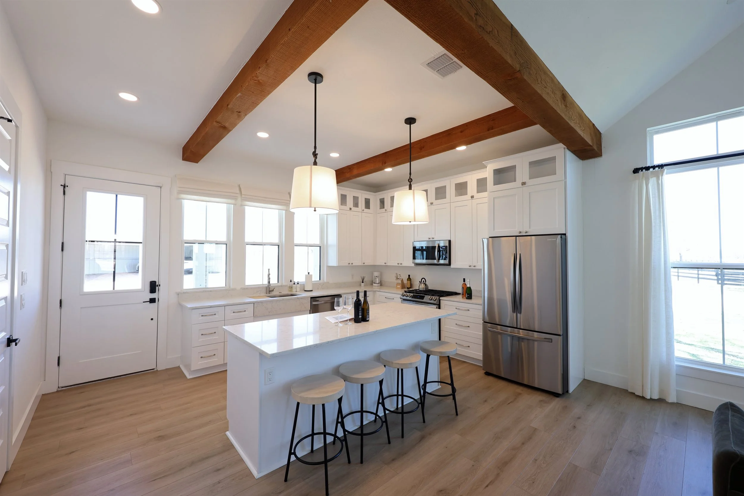 Modern kitchen with white cabinets, stainless steel refrigerator, island with seating, wooden beams on ceiling, pendant lights, and large windows.