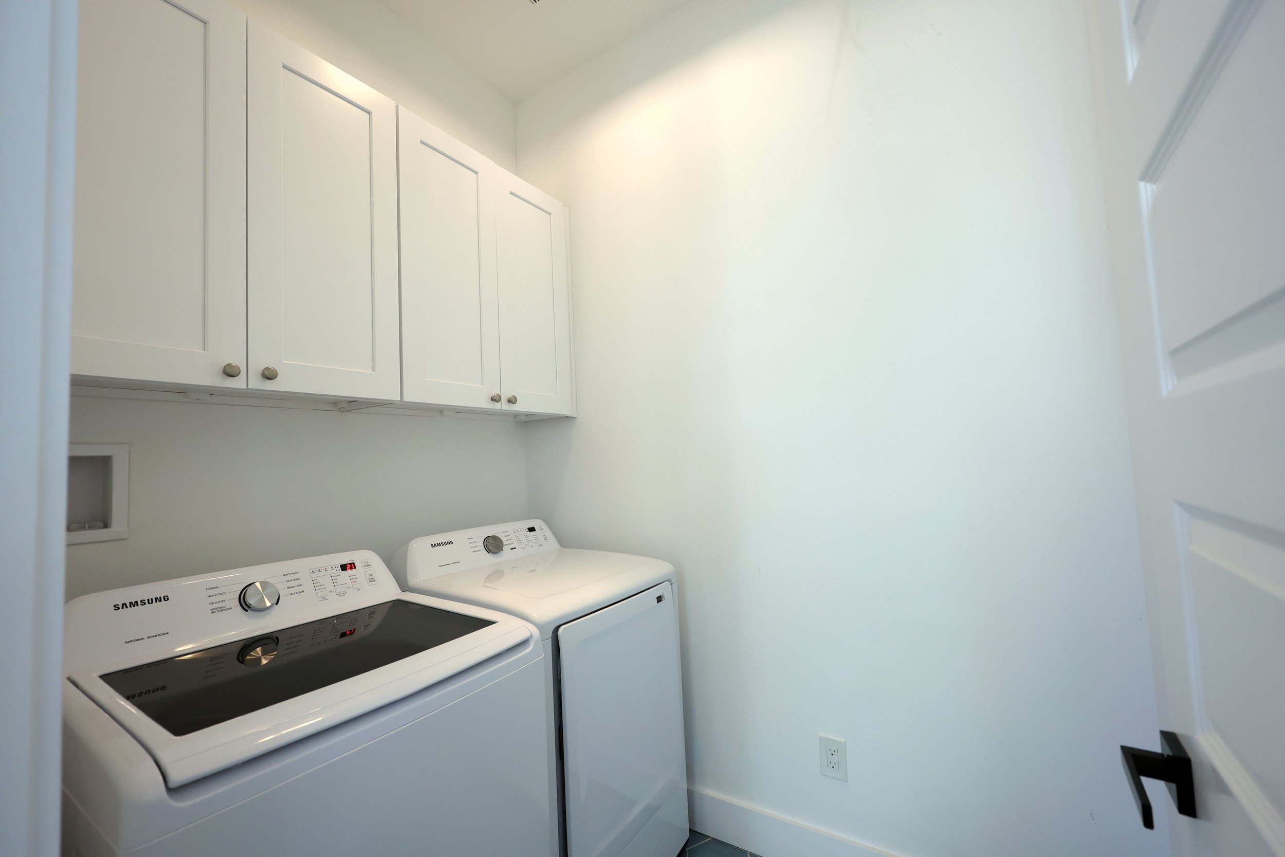A small laundry room with white cabinets above a white Samsung washer and dryer stack. The walls are painted light yellow, and there is an electrical outlet near the floor.