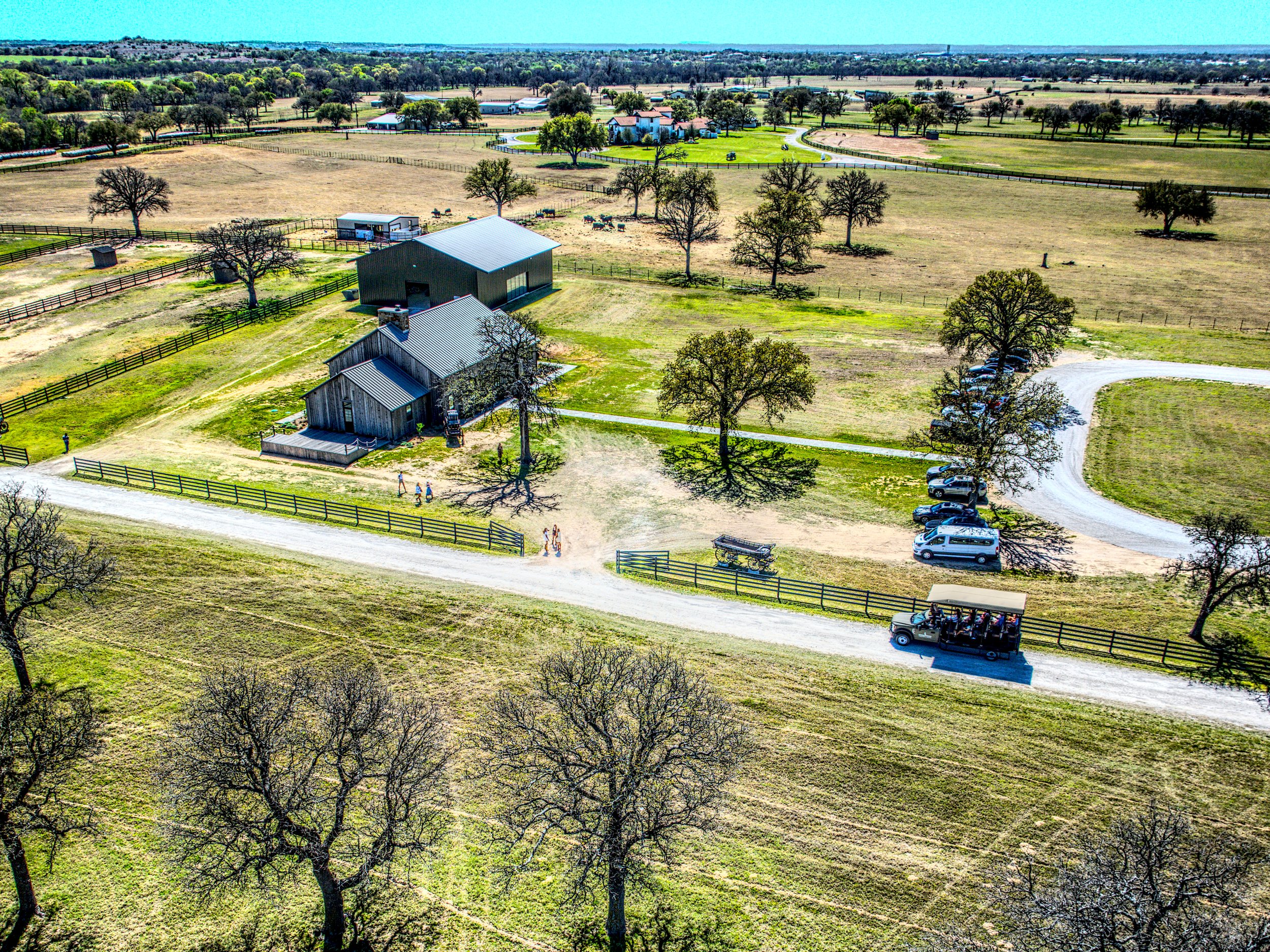 Aerial view of a ranch with several buildings, trees, fenced pastures, and parked cars on a dirt road under a clear sky.