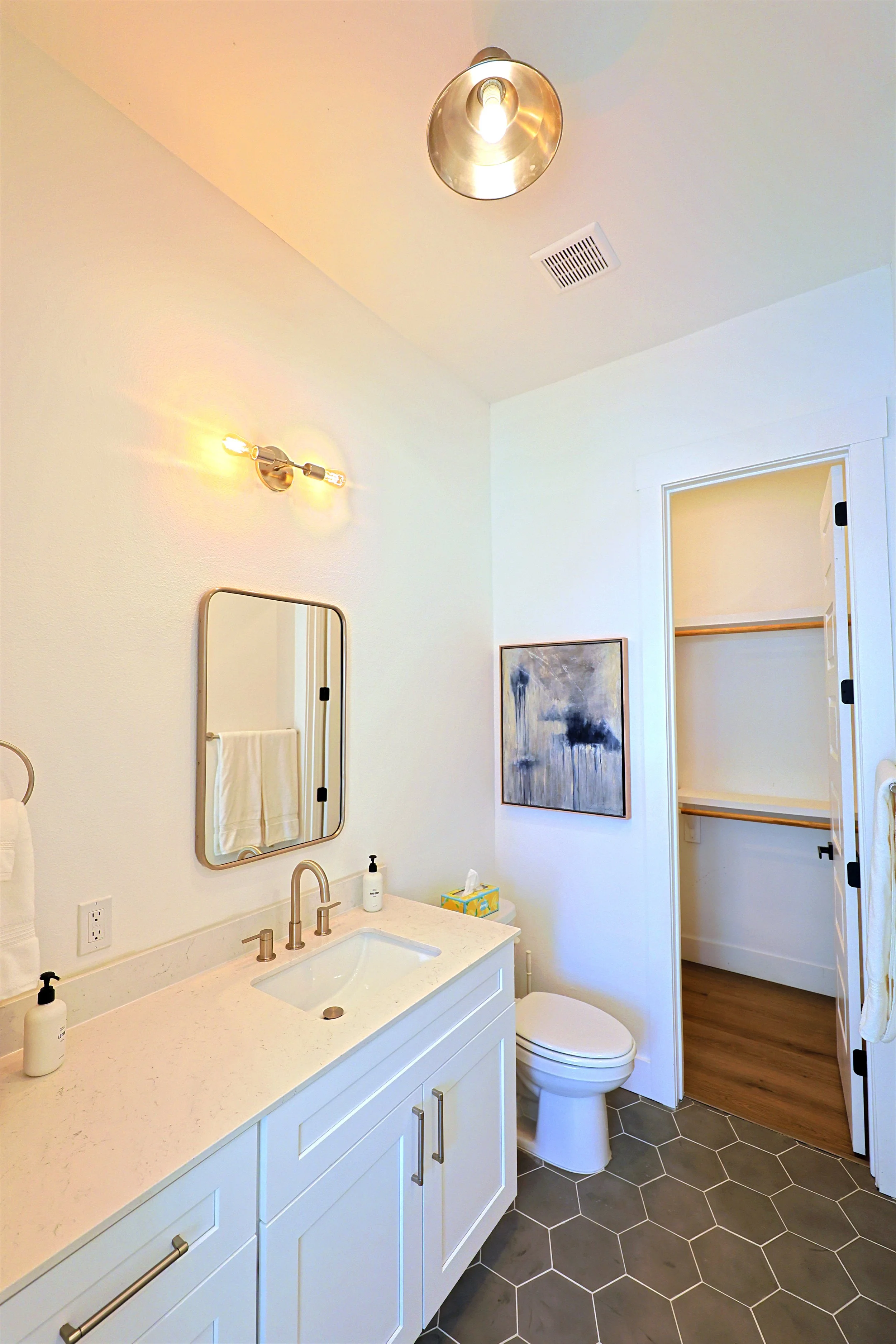 A bathroom with a white vanity, a mirror, a soap dispenser, a box of tissues, and a sink. There is a toilet and a storage closet with open shelves. The floor has gray hexagonal tiles, and the walls are painted white. There is a ceiling light and a wa