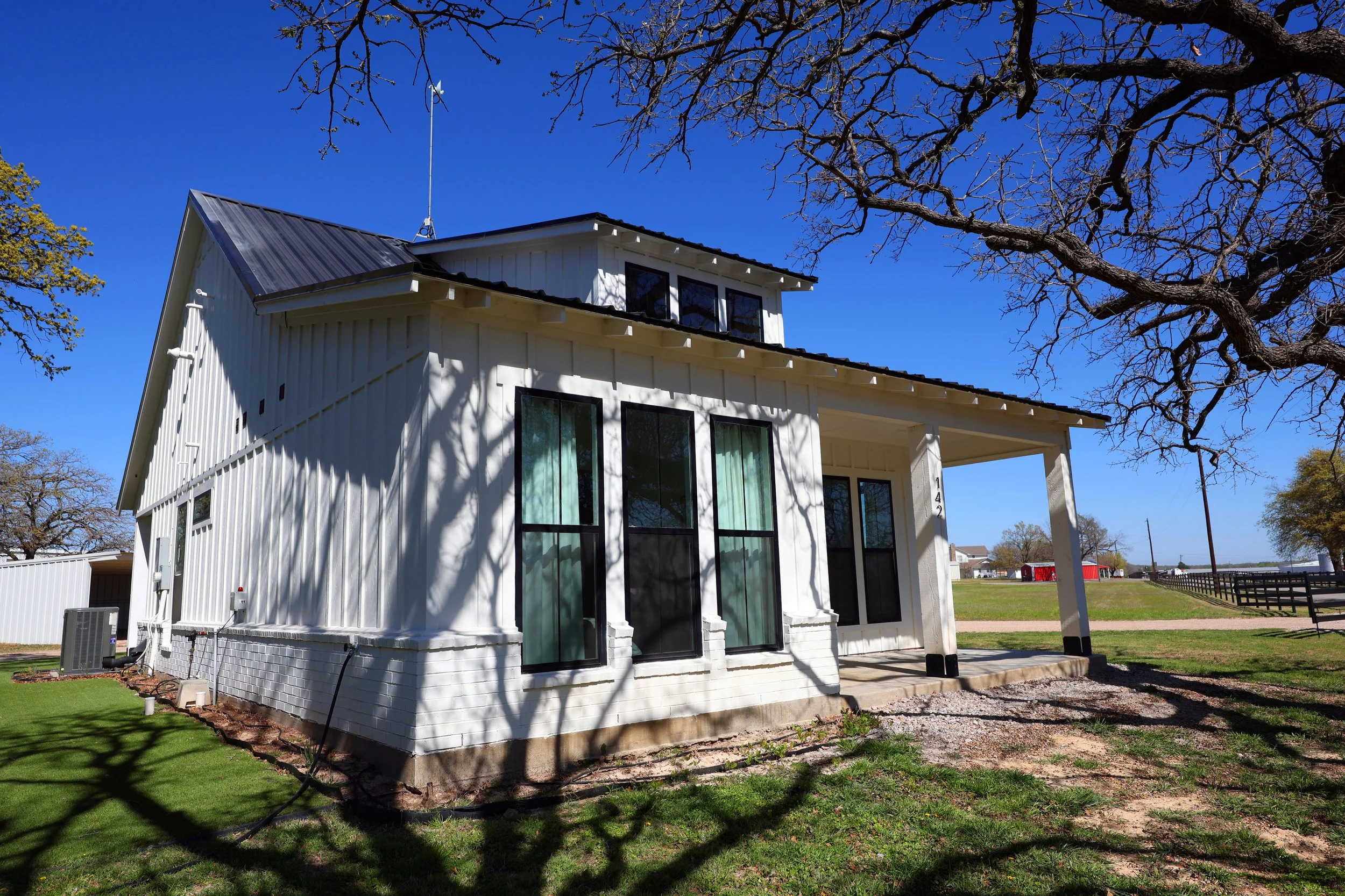White house with black trim and large windows, with a small porch and steps leading up. There are trees with shadows cast on the house, and the sky is clear and blue.