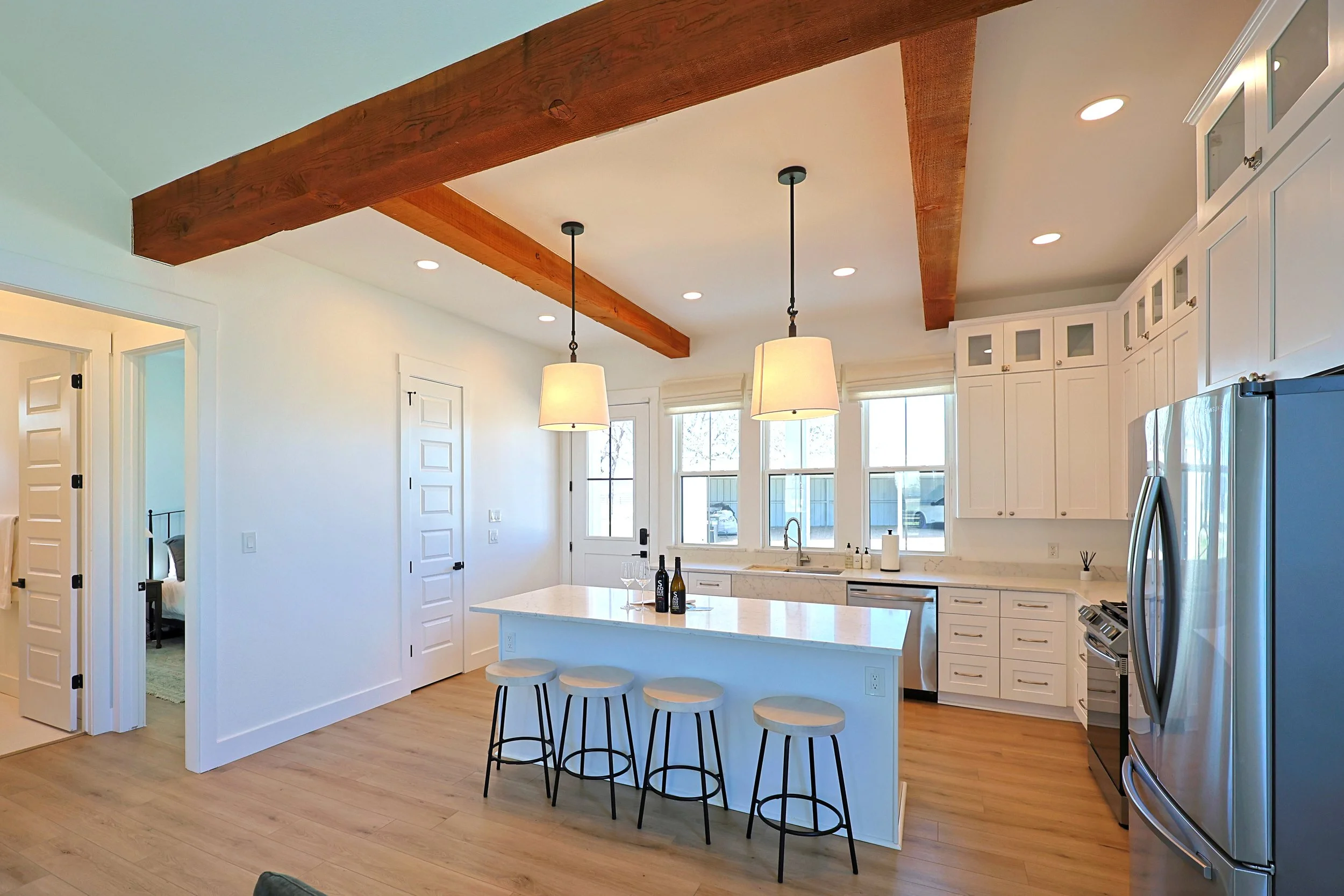 Modern kitchen with white cabinets, marble countertops, two hanging pendant lights, large windows, stainless steel refrigerator, and bar stools at an island.