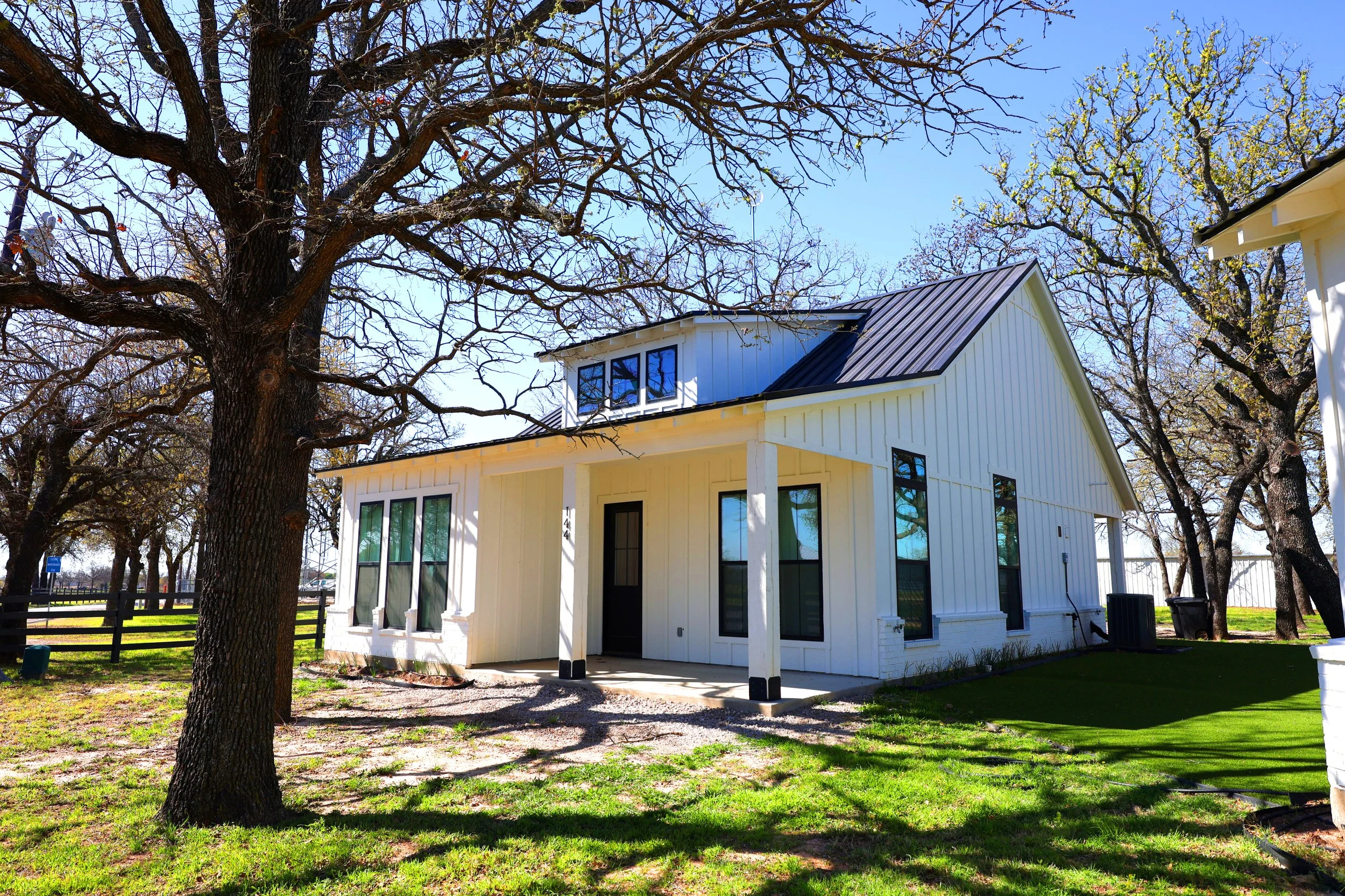 White modern house with black window frames and a metal roof, surrounded by leafless trees and a grassy yard under a clear blue sky.