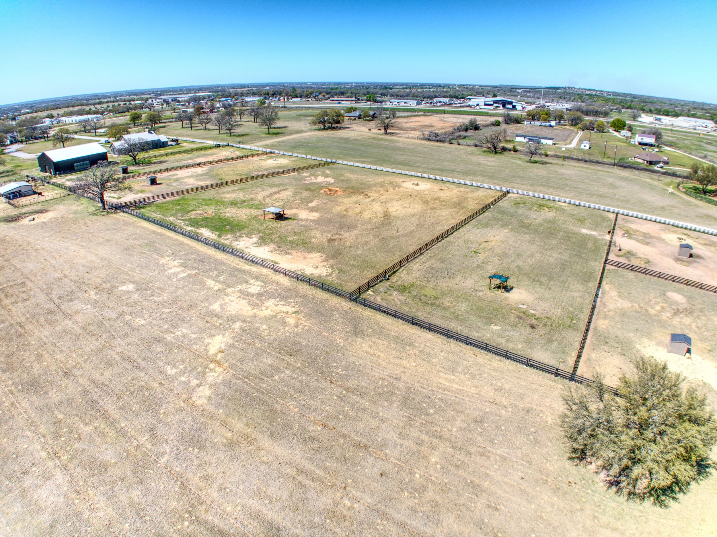 A wide aerial view of open fields, fenced areas, trees, and some buildings in a rural setting under a clear blue sky.