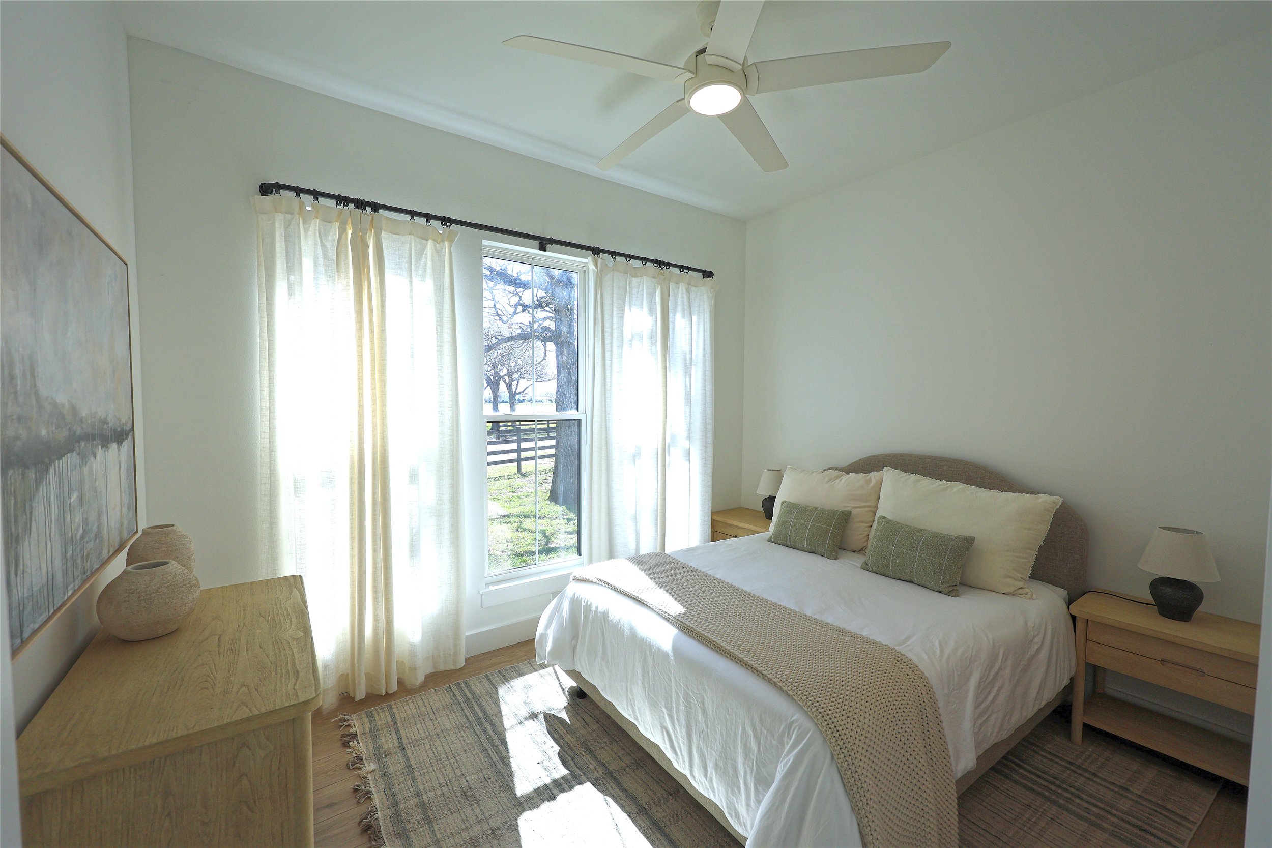 Bright bedroom with double bed, beige headboard, white bedding, and green pillows, next to two wooden nightstands with lamps, large window with sheer curtains overlooking a yard with a tree, ceiling fan with lights, and a painting on the wall.