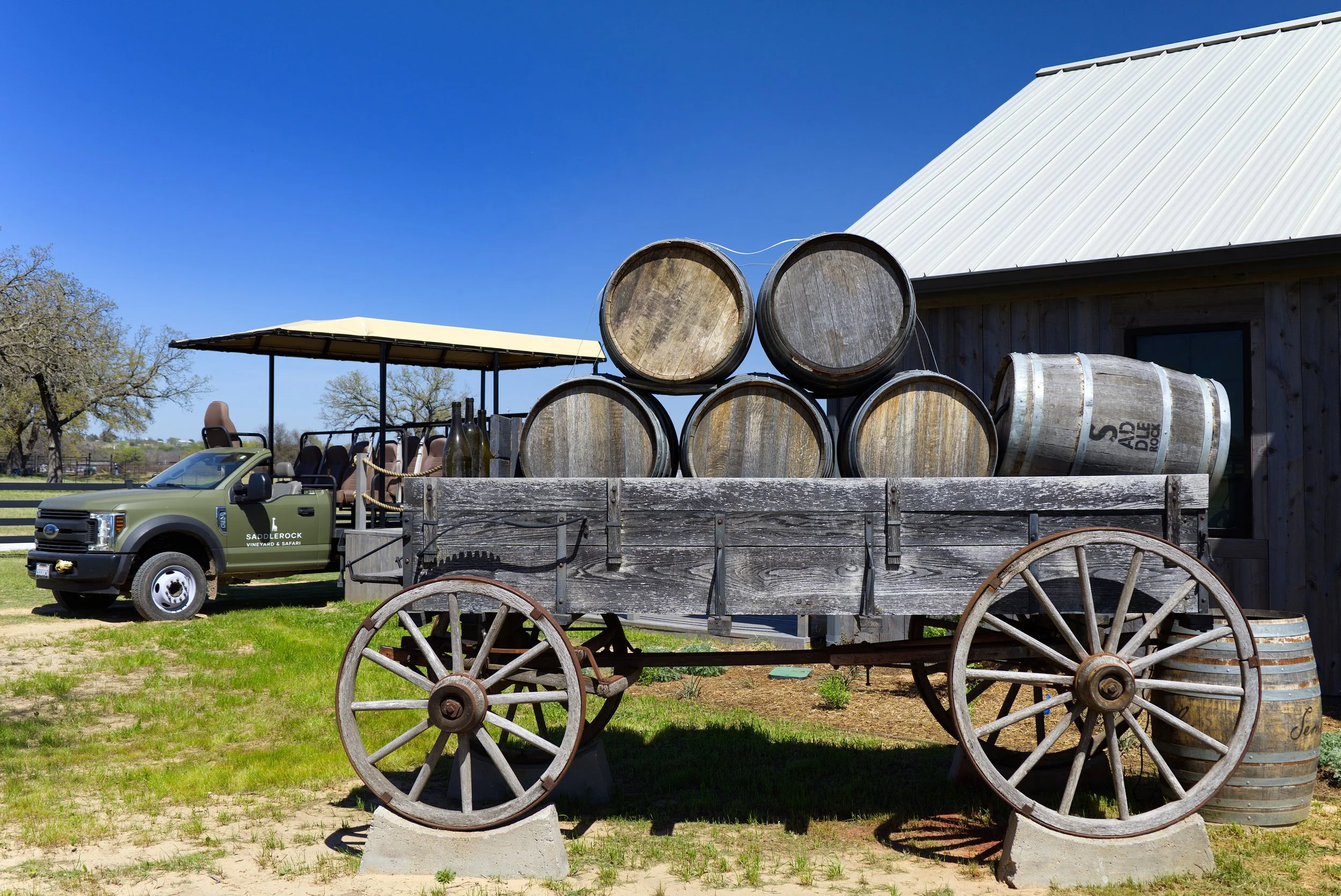 An old wooden wagon with large wheels and a wooden barrel on top. Behind it, there are stacked wine barrels, a building with a white metal roof, and a vehicle with a golf cart covered by a yellow canopy. The scene is outdoors with a clear blue sky an