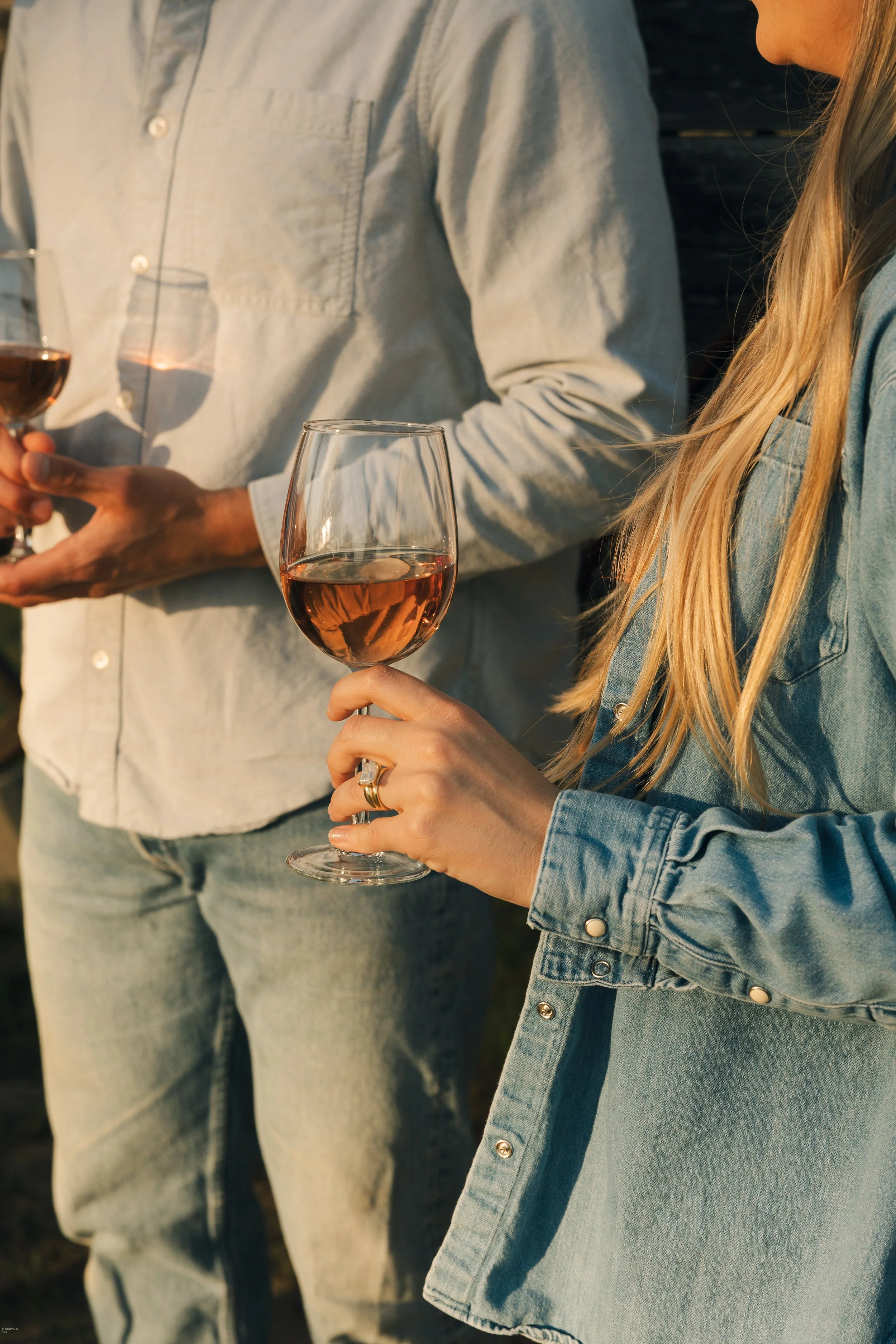A close-up of two people holding wine glasses filled with rosé wine, with only part of their bodies visible. One person is wearing a light-colored shirt, and the other is in a denim jacket.
