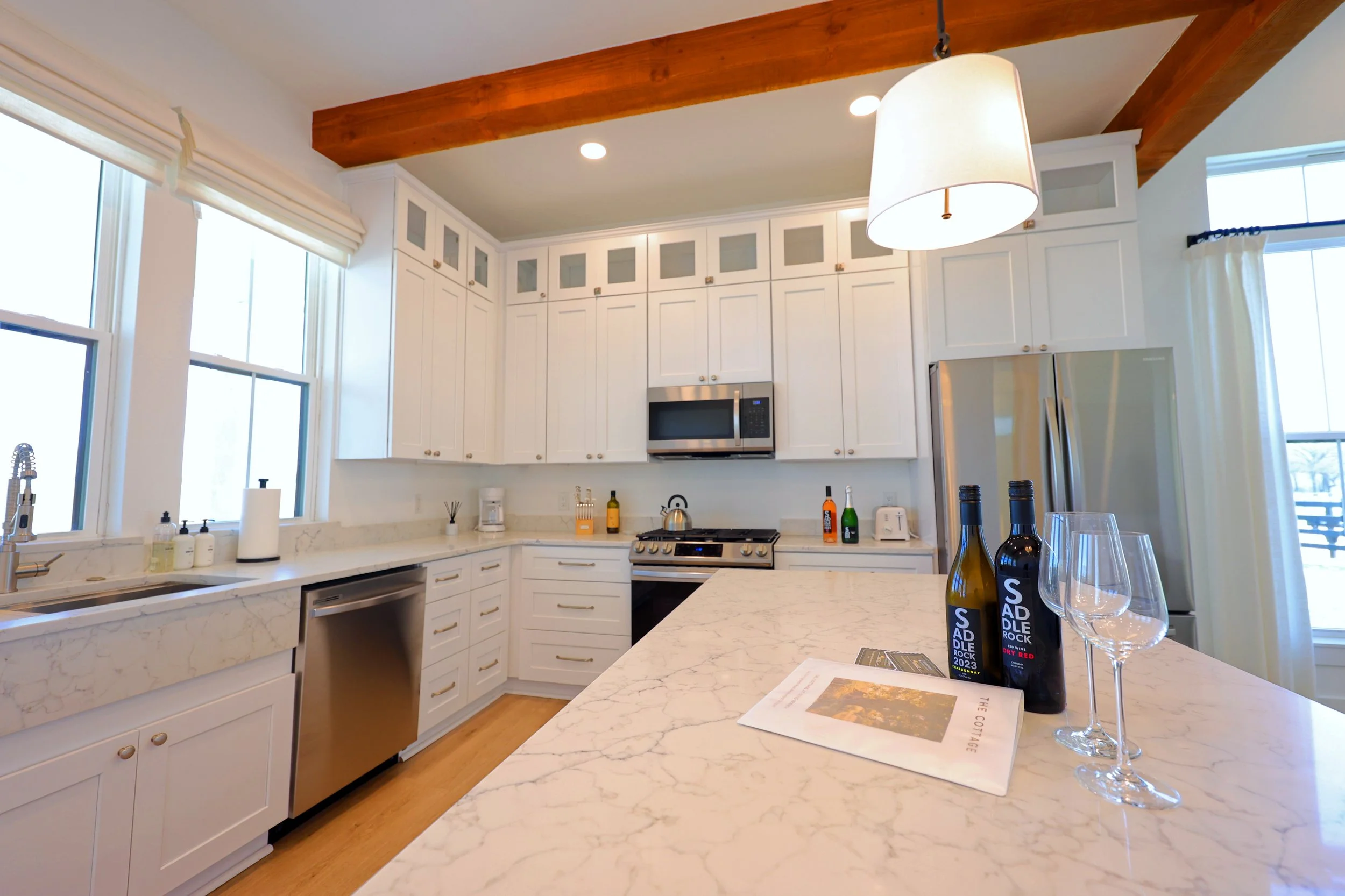 Modern kitchen with white cabinets, marble countertops, stainless steel appliances, and a wooden beamed ceiling. Items on the countertop include bottles of wine, wine glasses, paper towels, and small kitchen utensils. Large windows bring in natural l