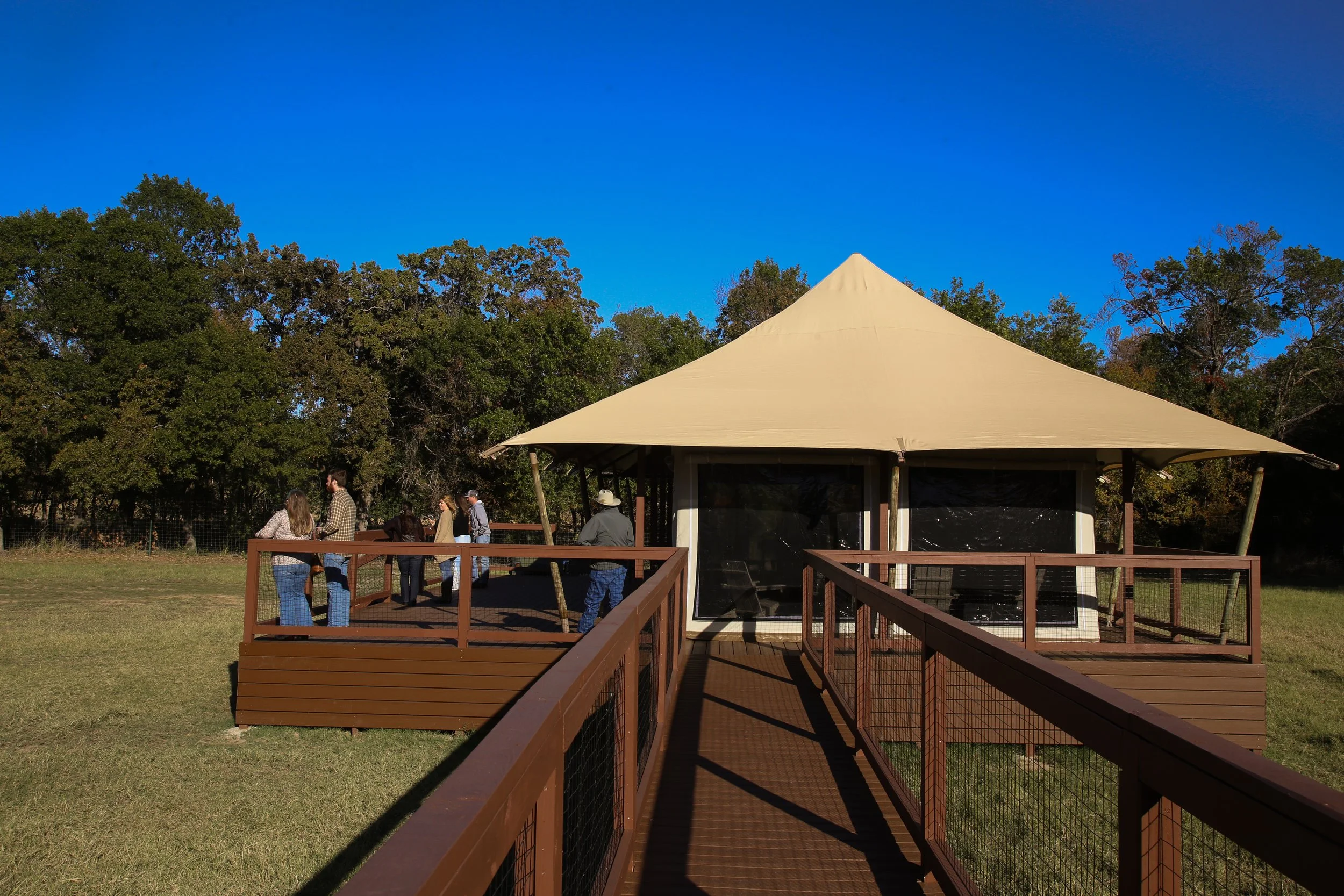 People gathering near a wooden deck and gazebo outdoors, with a blue sky and trees in the background.