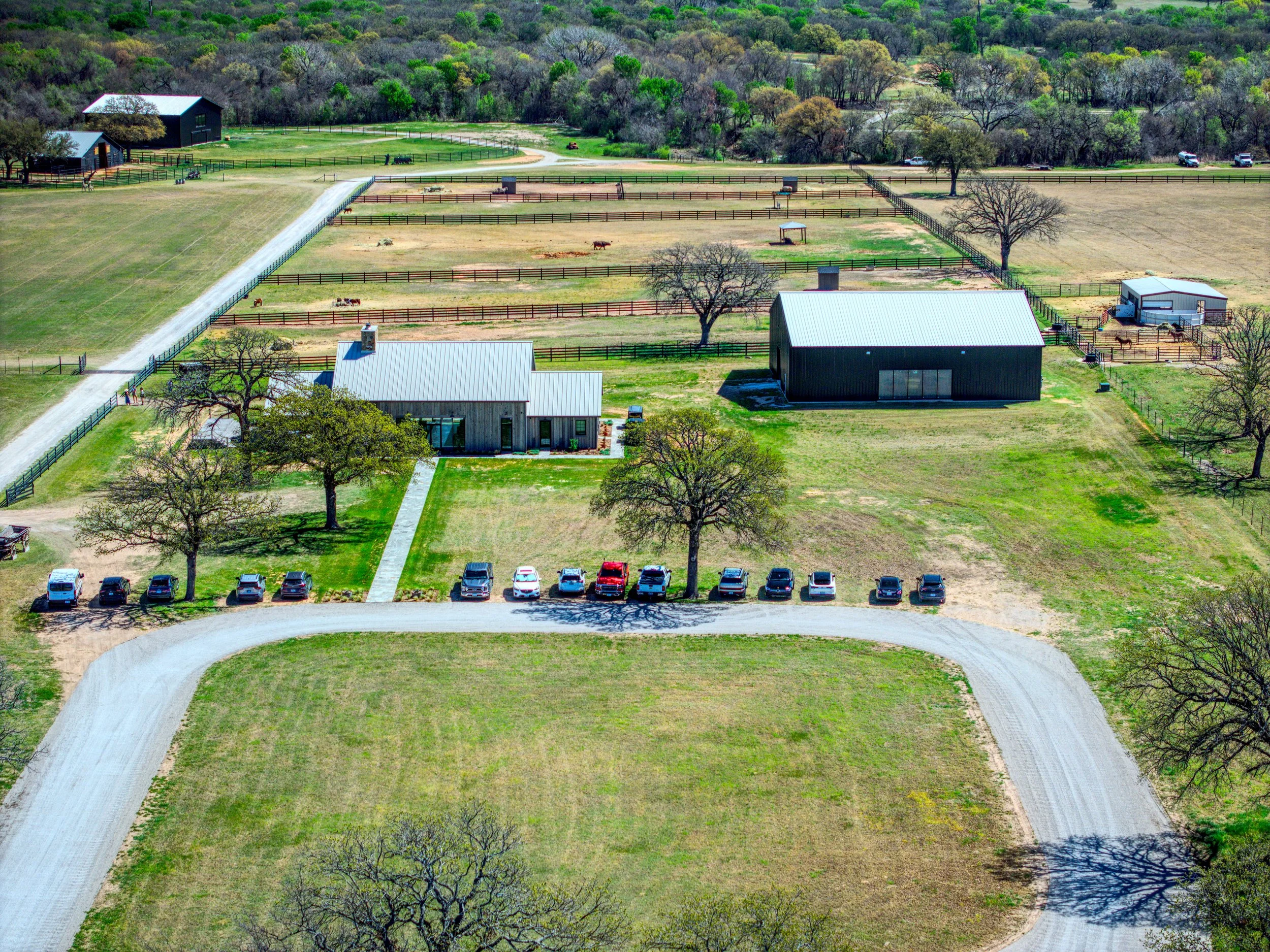 Aerial view of a farm with fenced animal enclosures, barns, trees, a gravel driveway, and parked cars, surrounded by green fields and wooded areas in the background.