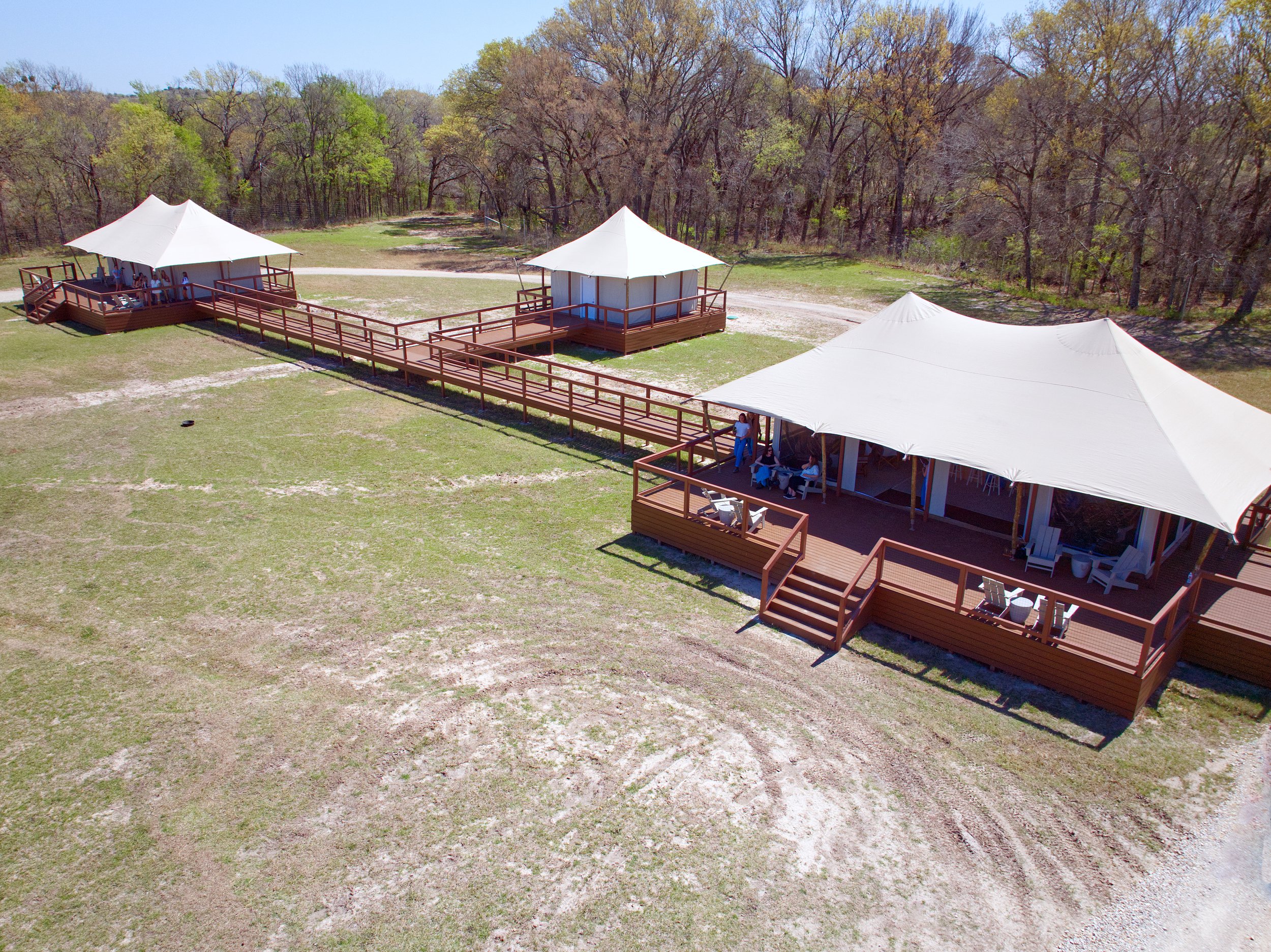 An outdoor scene with three white tents set up on a grassy area, connected by wooden walkways, with a wooded background.