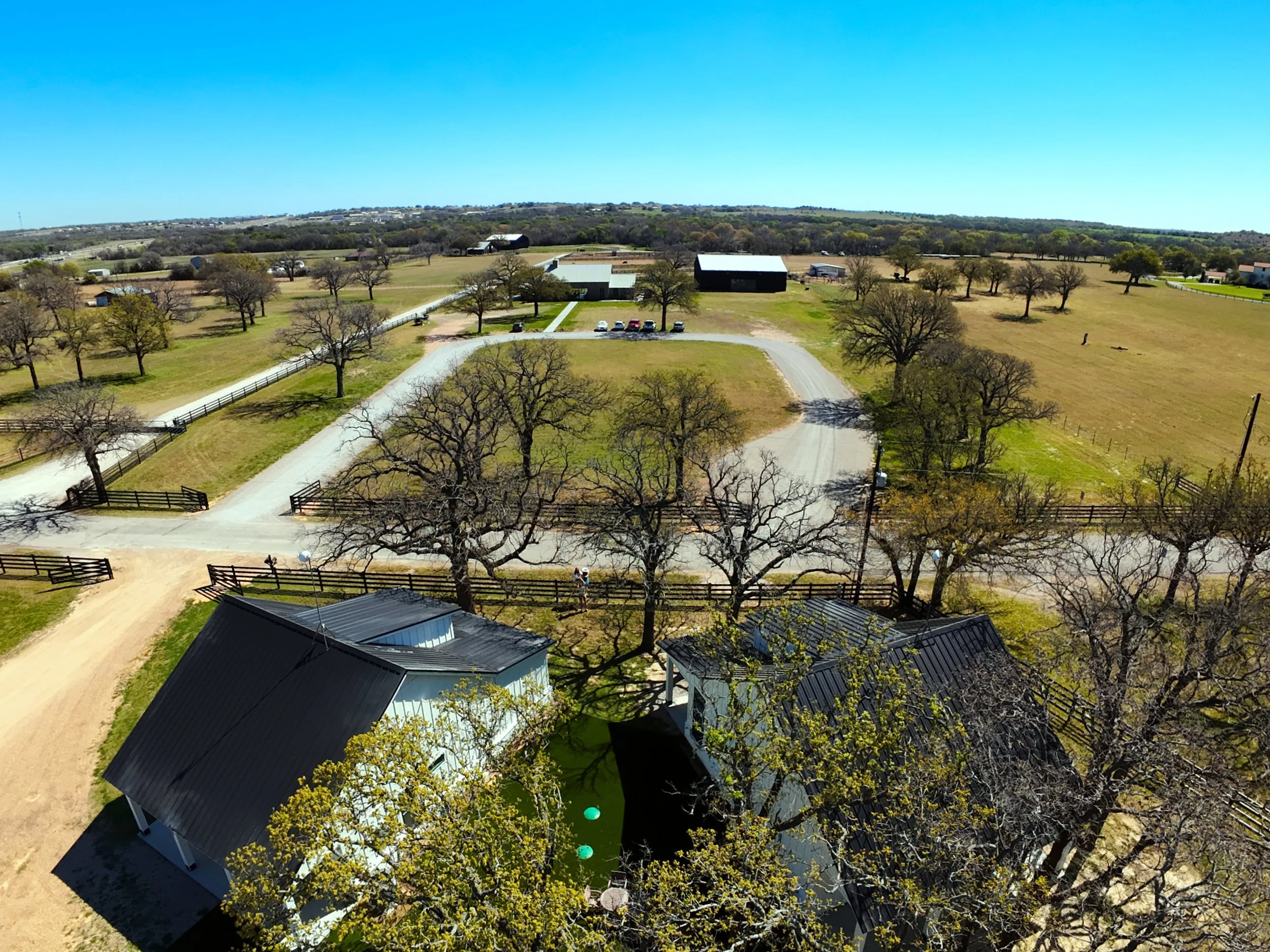 Aerial view of a countryside area with trees, farm buildings, a dirt driveway, and cars parked near a large barn, under a clear blue sky.