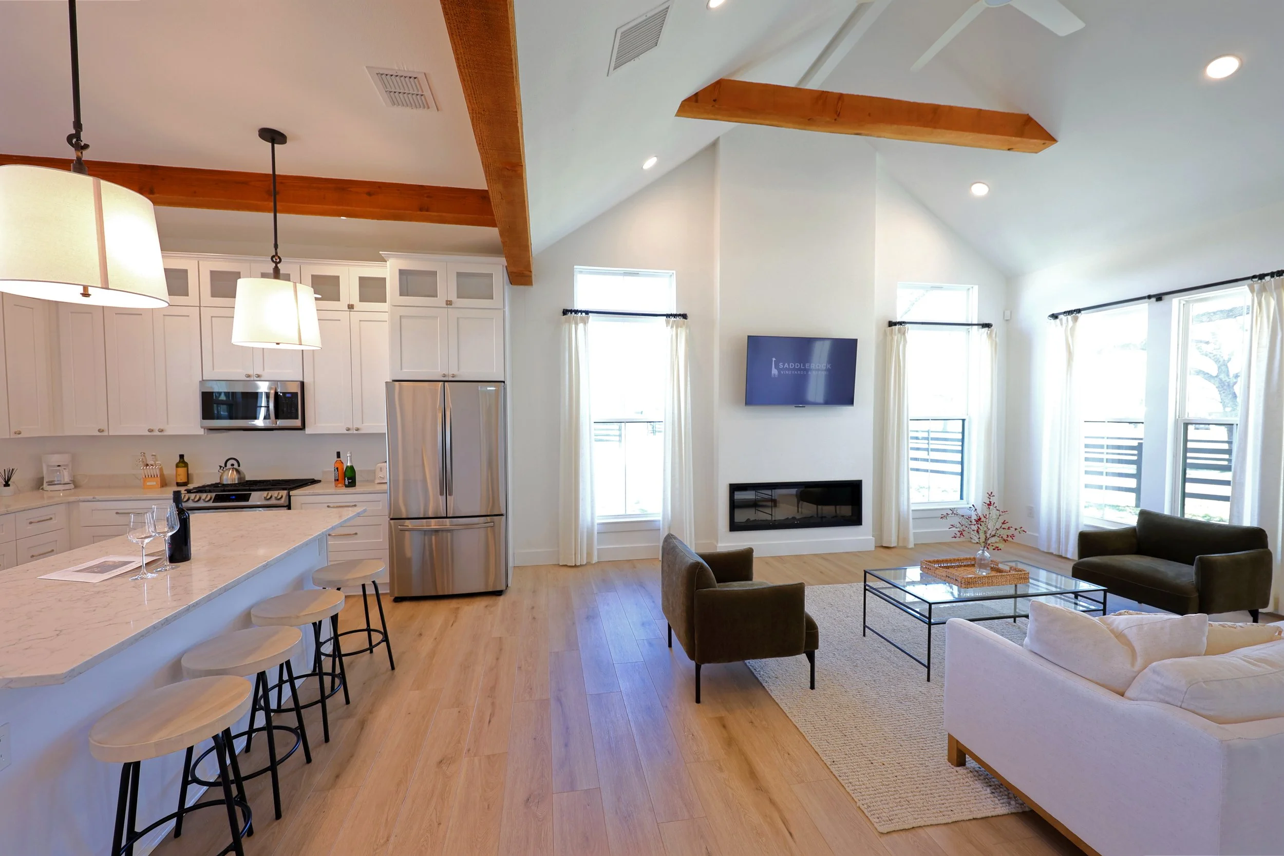 Open concept living room and kitchen with vaulted ceilings, exposed wooden beams, and large windows with white curtains, featuring modern furniture including a sofa, armchairs, and a glass coffee table, with a mounted TV above a fireplace.