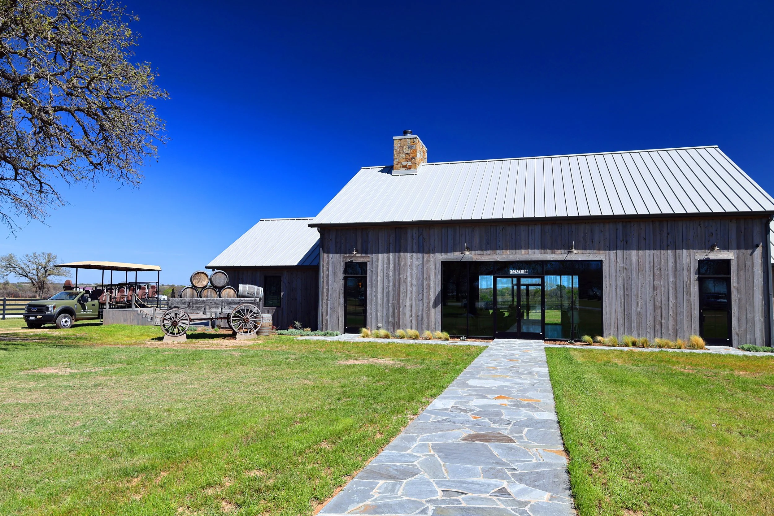 Exterior of a rustic barn with a metal roof, stone chimney, and large glass doors, surrounded by a grassy lawn with a stone pathway leading to the entrance, and a vintage wagon and barrels nearby, under a clear blue sky.