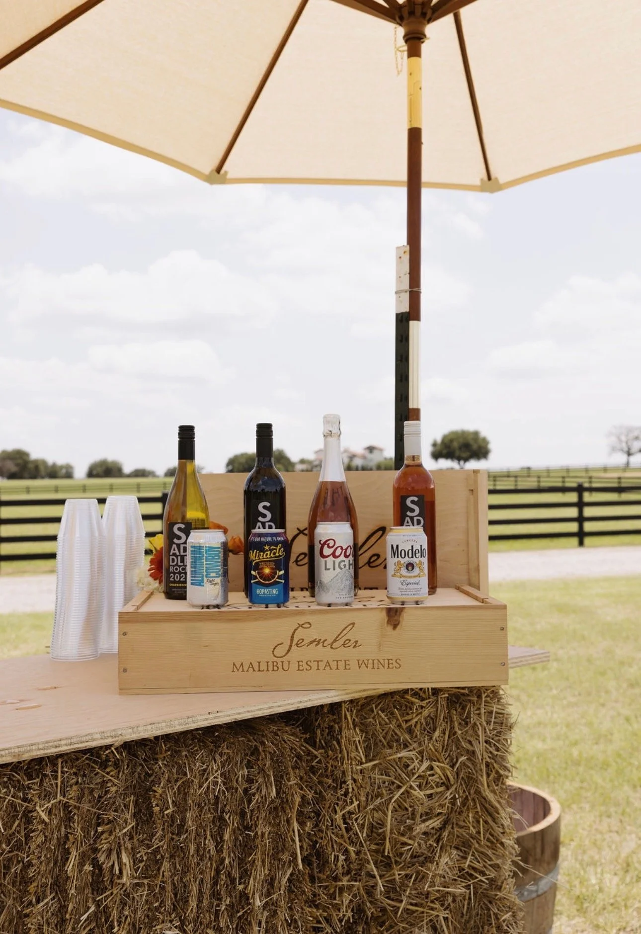 A wooden box labeled 'Semler Malibu Estate Wines' with four bottles of wine, paper cups, and a can of soda on top, set outdoors under a large beach umbrella on a bright day with a field and trees in the background.