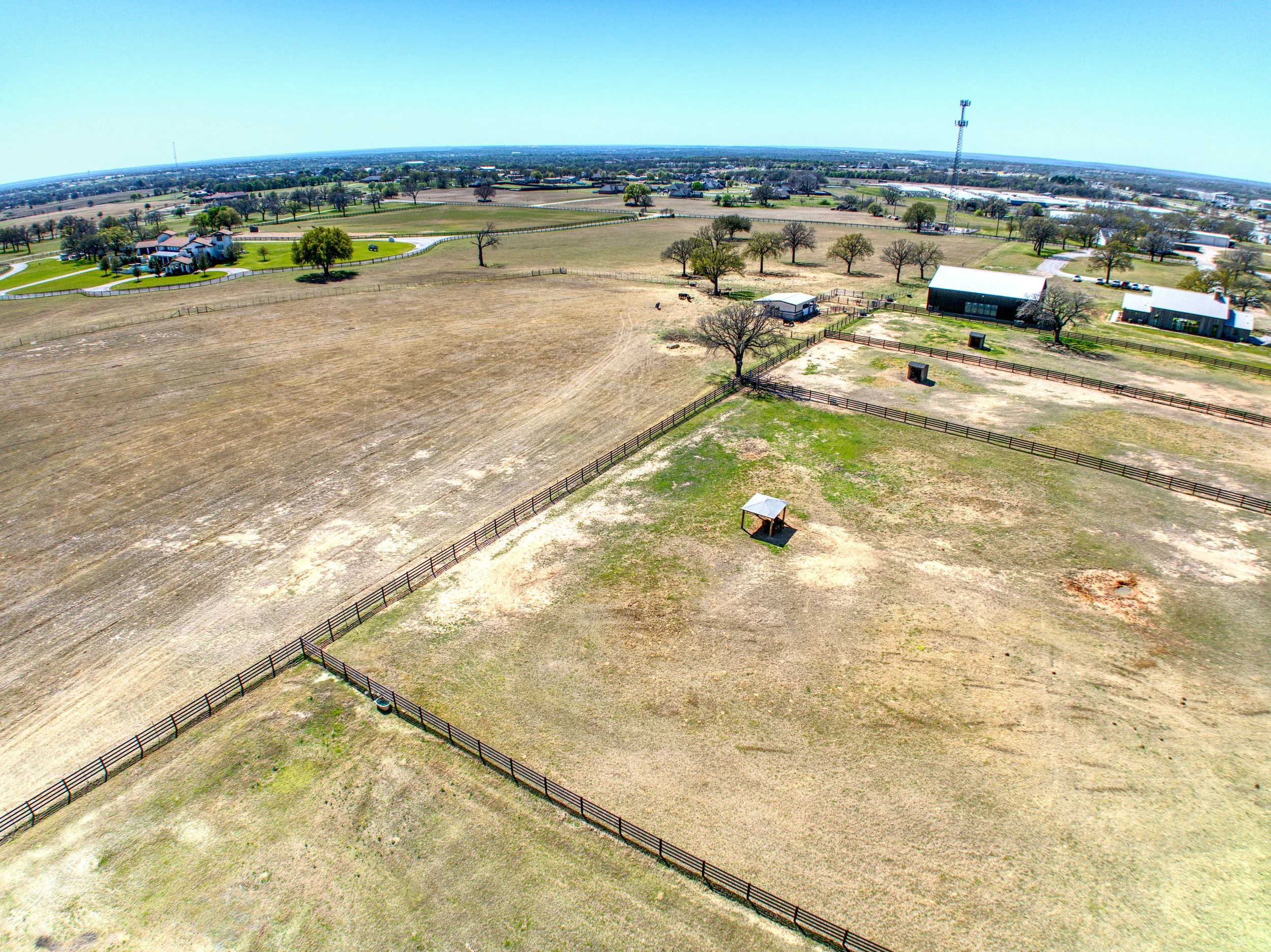 Aerial view of a rural landscape with fenced-in pastures, trees, a house, barns, and open fields under a clear blue sky.