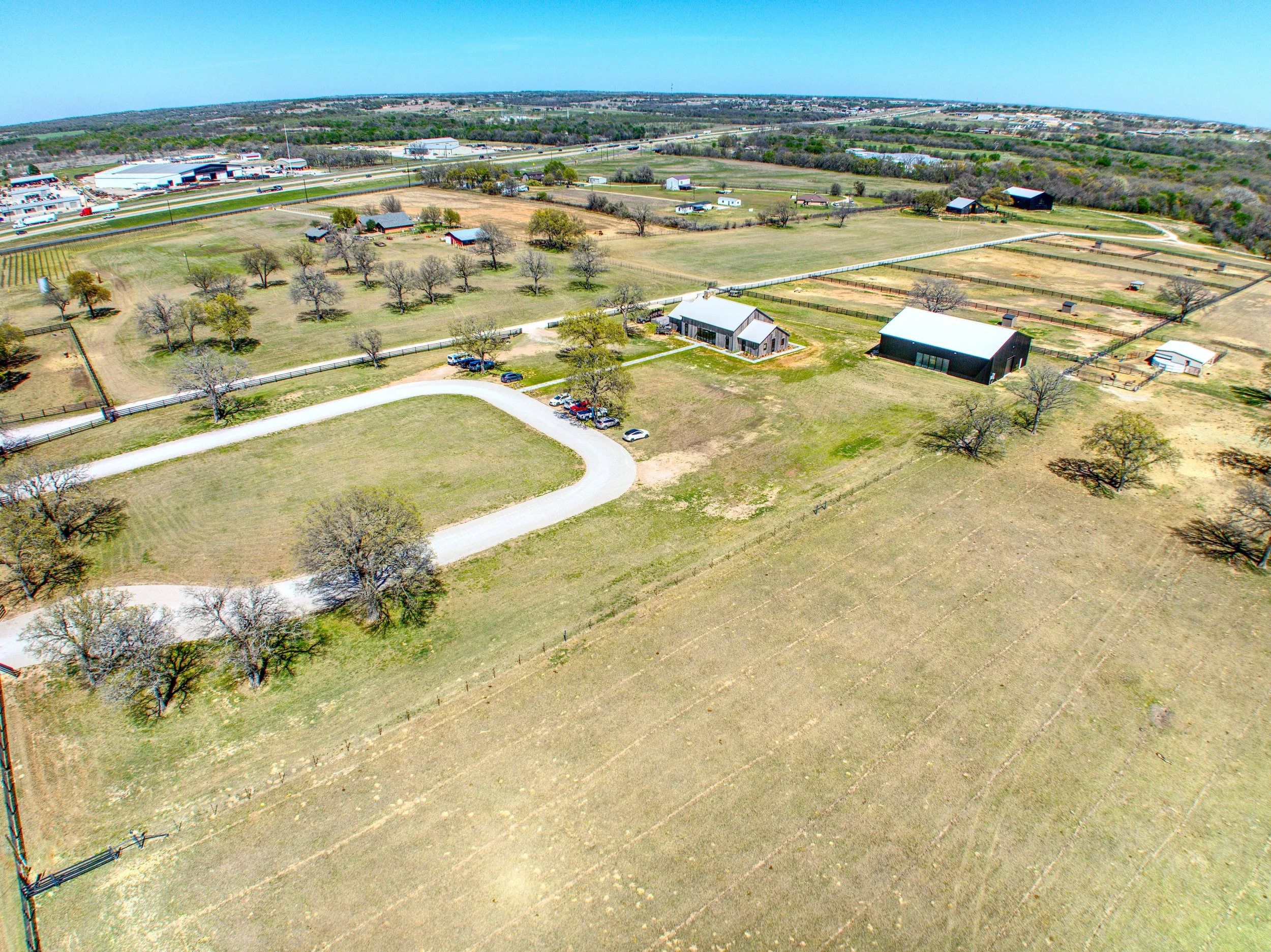 Aerial view of a rural farm with fenced fields, several barns, trees, a winding driveway, and a distant industrial area and highways.