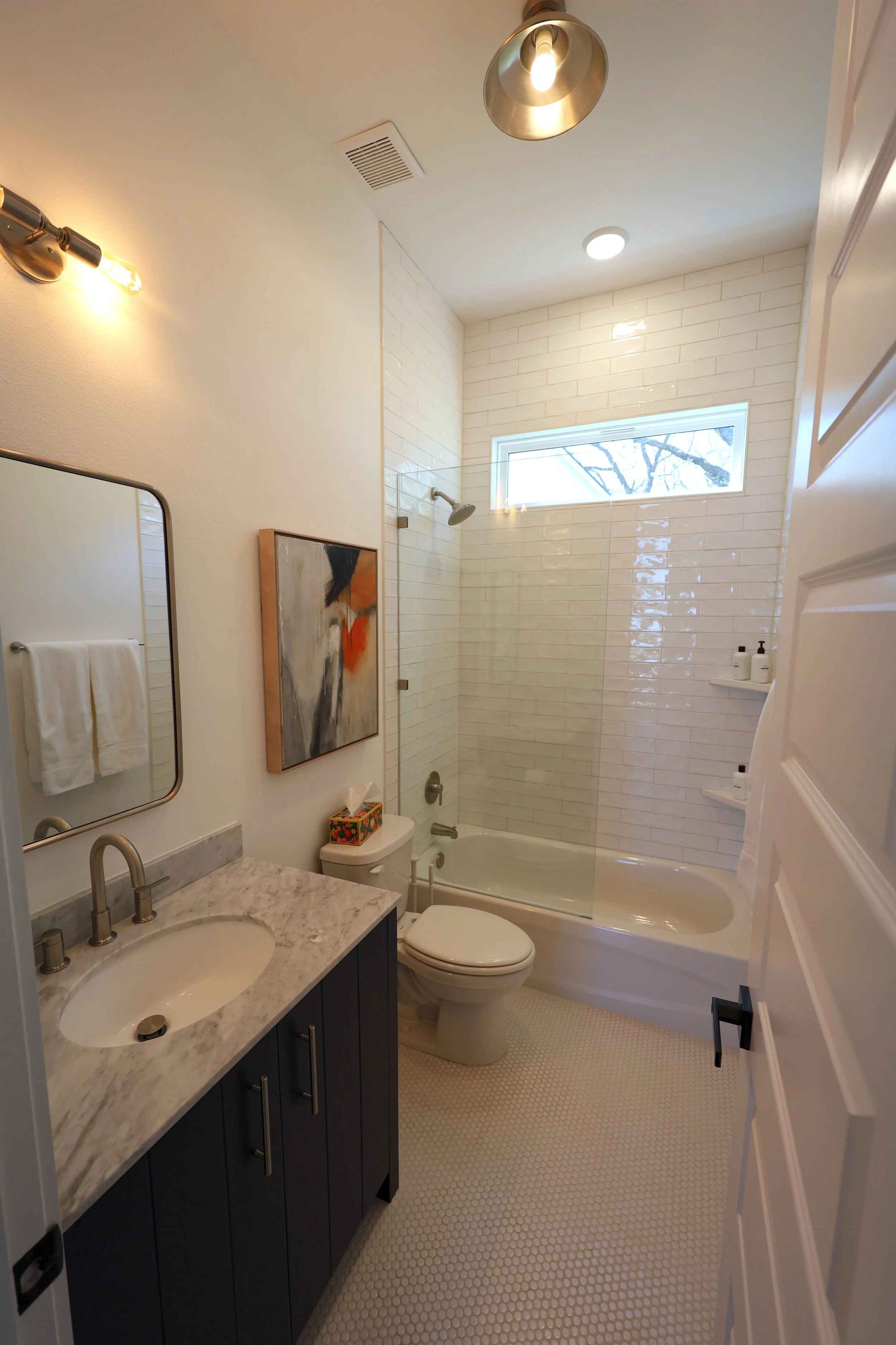A modern bathroom with a white bathtub and shower, a vanity with a marble countertop, a mirror, a framed abstract artwork, a toilet, and a window with a view of trees. The ceiling has a light fixture and a vent, and there are white tile walls and a t